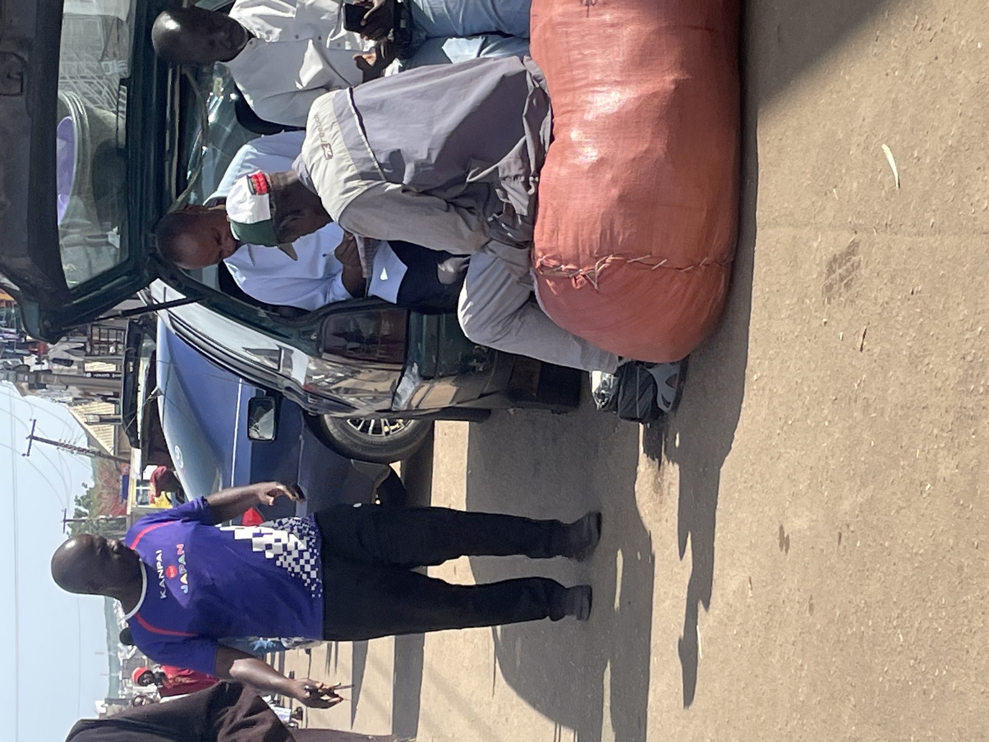 The image depicts a street scene in Jos, Nigeria, with a focus on a group of men gathered around the open trunk of a car. A man in a purple t-shirt with "KANPAI JAPAN" visible on it is walking towards the left of the frame. Inside the trunk of a dark green car, two men are seated. One man, wearing a white shirt, is looking down and appears to be writing on a piece of paper. Another man, wearing a light-colored jacket over a t-shirt and a cap, is seated on a large, reddish-orange cylindrical bag. A third man, wearing a white shirt, is partially visible to the right, holding a phone. The ground is dusty and paved. The background shows buildings, streetlights, and other vehicles, suggesting a busy urban environment. Shadows are cast on the ground, indicating the presence of sunlight.