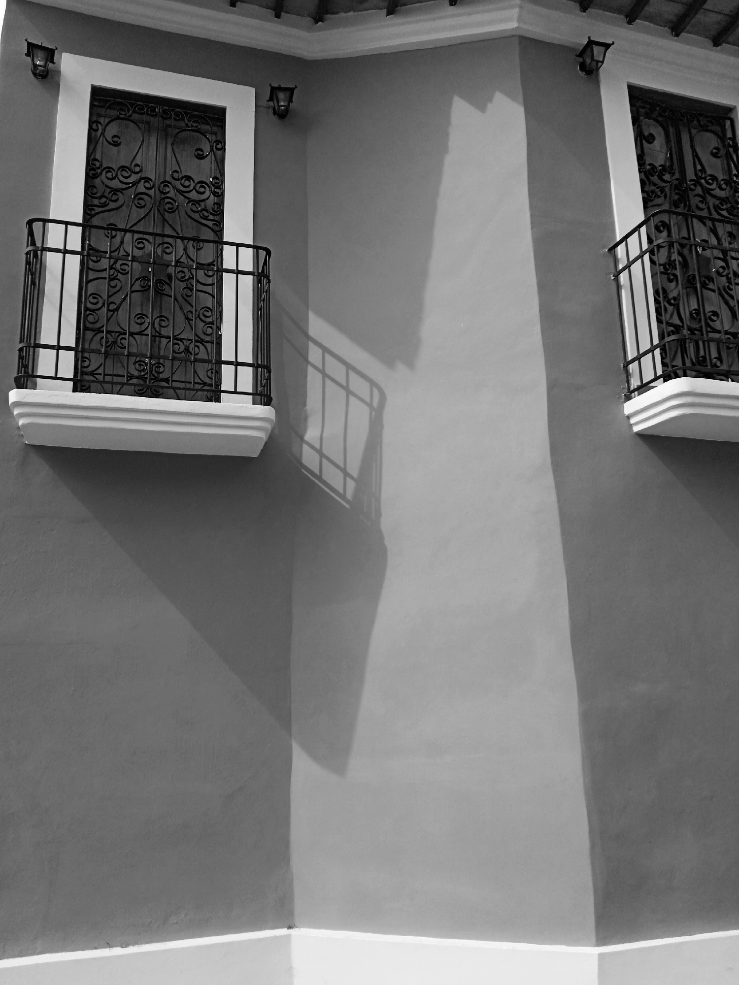 Black and white photograph showing two ornate wrought iron balconies on a building in Montalbán, Venezuela. The building's exterior wall is a uniform gray tone. The balconies have white trim. The lighting casts a distinct shadow from the left balcony onto the wall, creating a geometric shape. A white baseboard runs along the bottom of the building. There are two small light fixtures above each balcony.