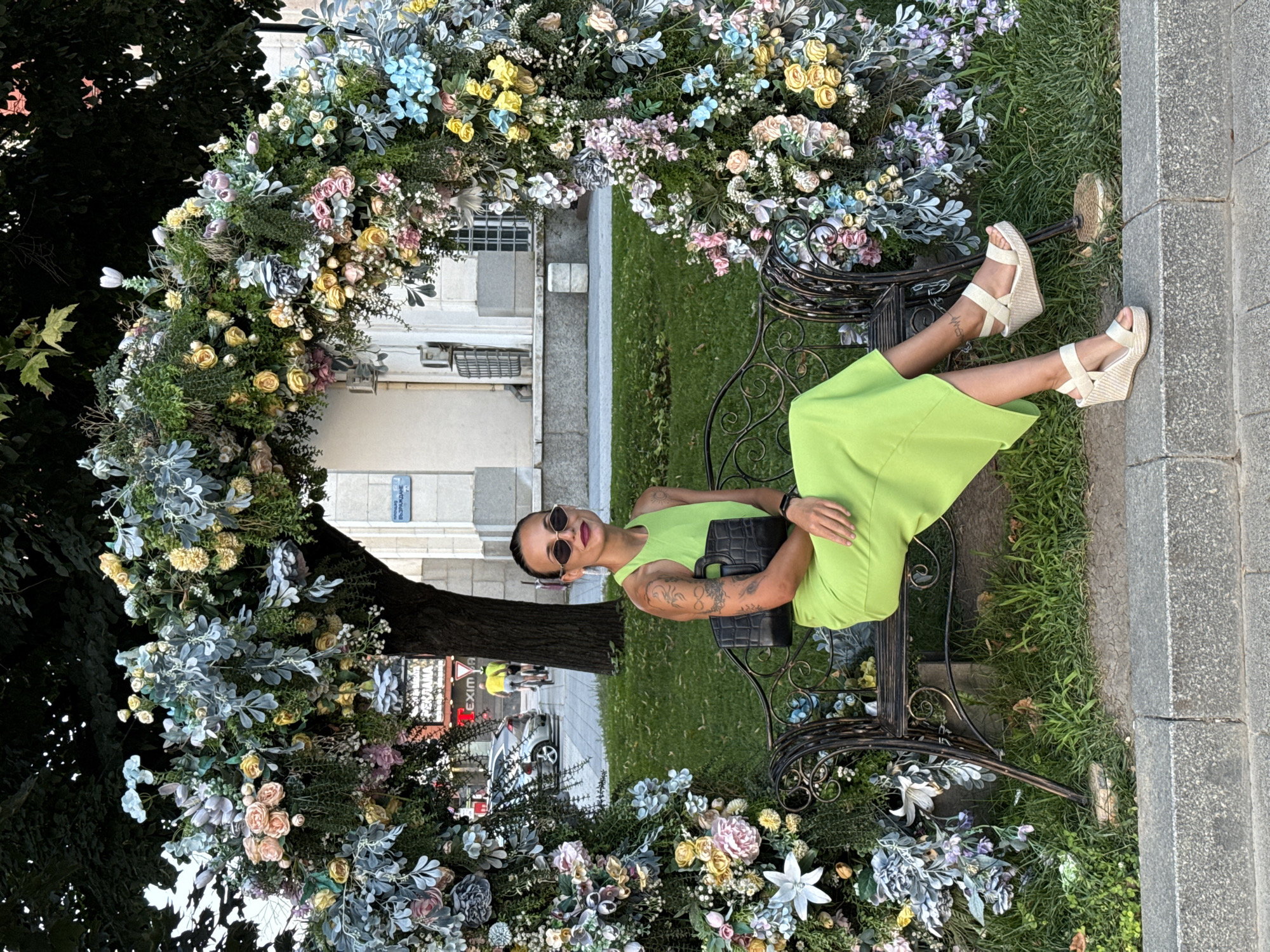 A woman sits on a wrought-iron bench under a floral archway. She wears a sleeveless lime green dress, sunglasses, and wedge sandals, and carries a black handbag. The arch is adorned with an assortment of artificial flowers and greenery, including pink, yellow, blue, and white blossoms. Behind the arch, a tree trunk is visible, and beyond that, a building with arched windows. In the background, a street with cars and cyclists can be seen, suggesting an urban setting. The bench is situated on a grassy area adjacent to a paved walkway. This scene is located in Pleven, Bulgaria, specifically in front of the Regional Historical Museum "Parashkev Tsvetkov".