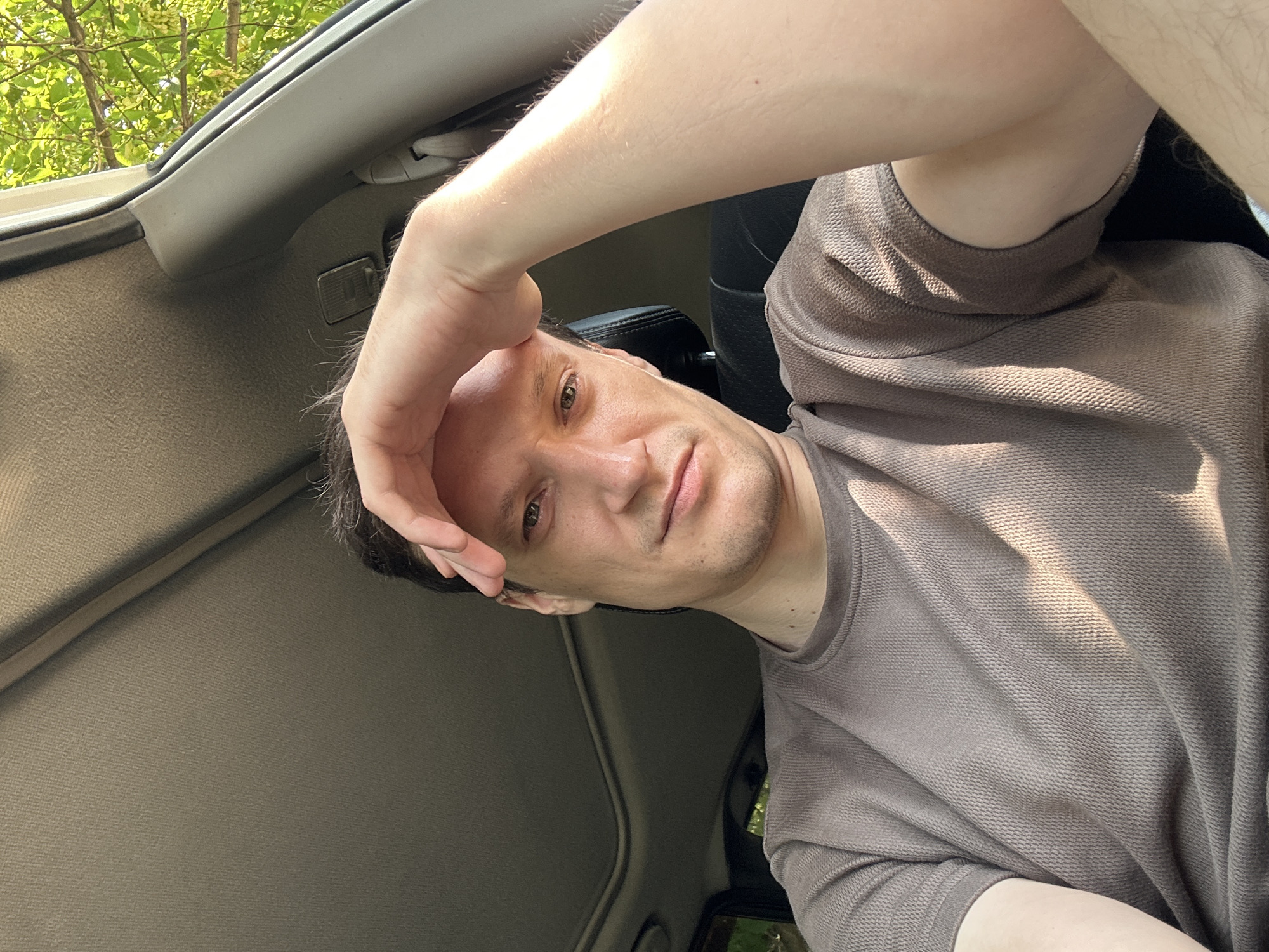 A man is sitting inside a car, seen from a low-angle, selfie-style perspective. He is wearing a brown, textured t-shirt. His right hand is resting on his forehead, and he is looking towards the camera with a neutral expression. Sunlight is streaming into the car from the right, casting shadows across his face and shirt. The interior of the car is visible, including the ceiling with a sunroof. Outside the car, through the open window on the right, green foliage from trees can be seen. The image appears to have been taken during daylight hours. The exact location within the city of Makiv, Ukraine cannot be determined from the image alone.