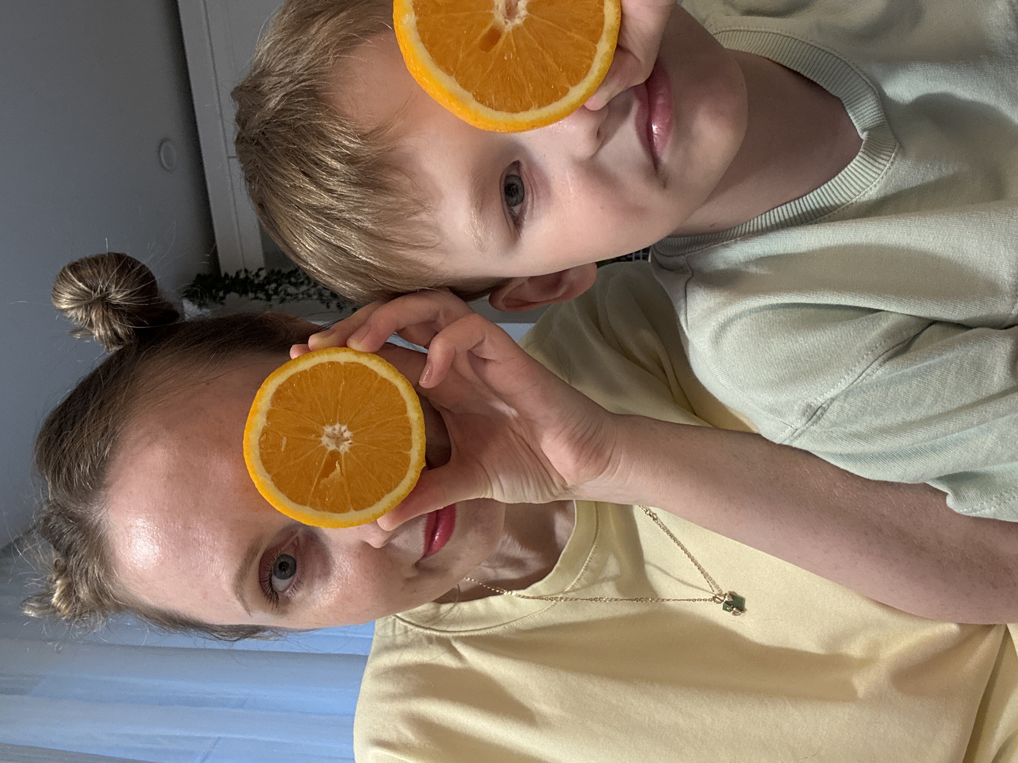 A woman and a child are posing with orange slices in front of their eyes. The woman has light hair pulled into a bun, a yellow t-shirt, and a gold necklace. The child has short blonde hair and a light green t-shirt. The background is blurry but appears to be indoors with a light blue wall visible. The location is Chaikovsky, Russia.
