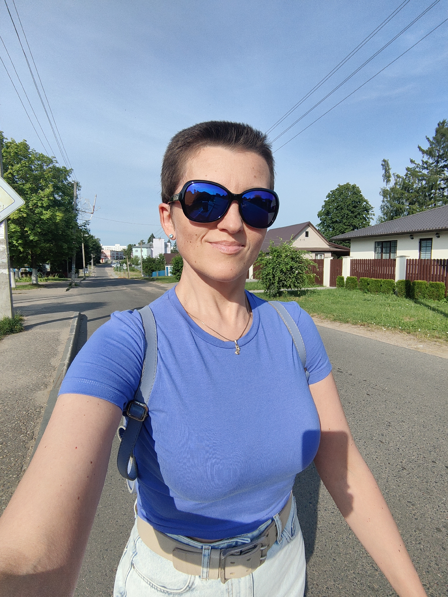 A woman with a short buzz haircut and large, dark sunglasses is captured in a selfie on a paved road in Fanipal, Belarus. She is wearing a vibrant blue t-shirt and light-colored denim shorts, cinched with a beige belt. A light blue backpack is slung over her left shoulder. Her right arm is extended, holding the camera, and her left arm is partially visible. The background features a suburban street with houses, green trees, and utility poles with overhead wires. The sun is bright, casting shadows on the road and the woman's arm. In the reflection of her sunglasses, the selfie-taker is visible along with the surrounding environment.