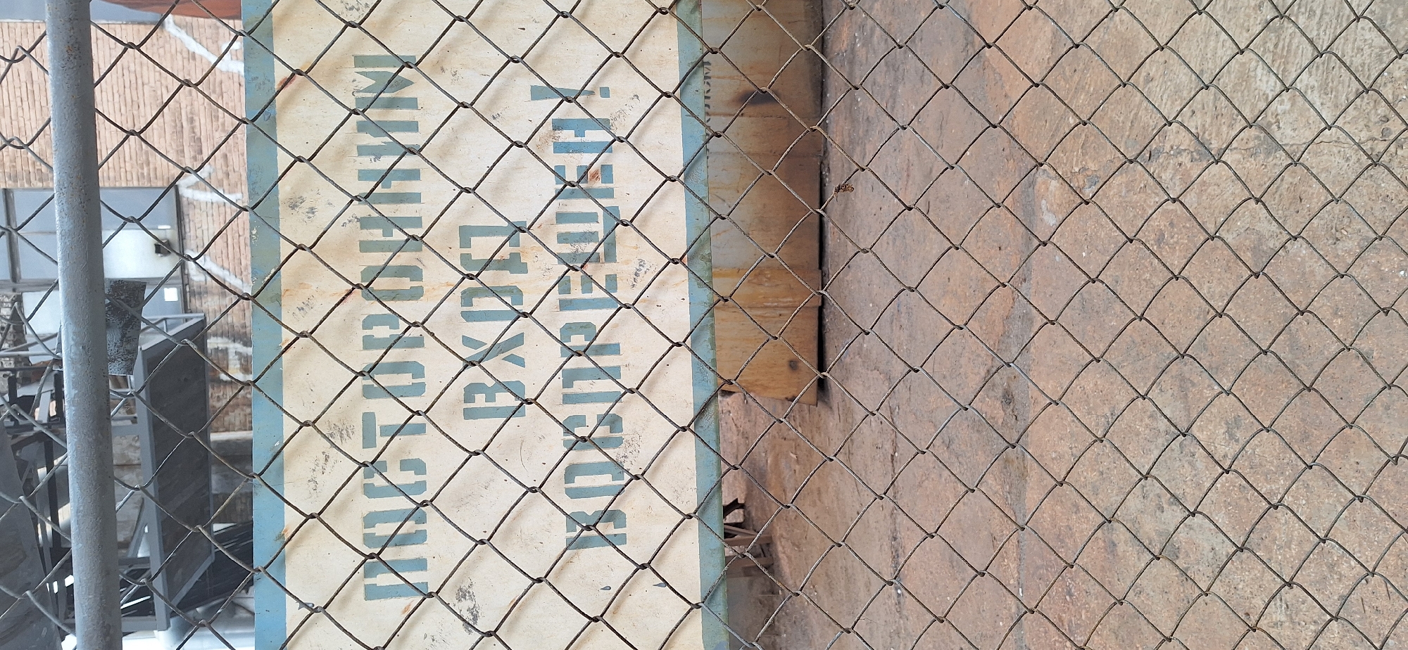 A metal chain-link fence is in the foreground of this image, obscuring a sign and a building. The sign, which is rectangular with a light-colored background and blue text, appears to be mounted on a wooden surface. The text on the sign is partially visible, reading "NATIONAL EXPORT". Behind the sign and fence, a brick building is visible. To the left of the sign, a vertical metal pole, likely part of the fence, is present. To the right of the sign, a textured, reddish-brown surface, possibly a wall or the ground, is visible. The scene appears to be outdoors, with natural lighting. There are no people visible in the image. The context provided indicates the location is Nakhodka, Russia.