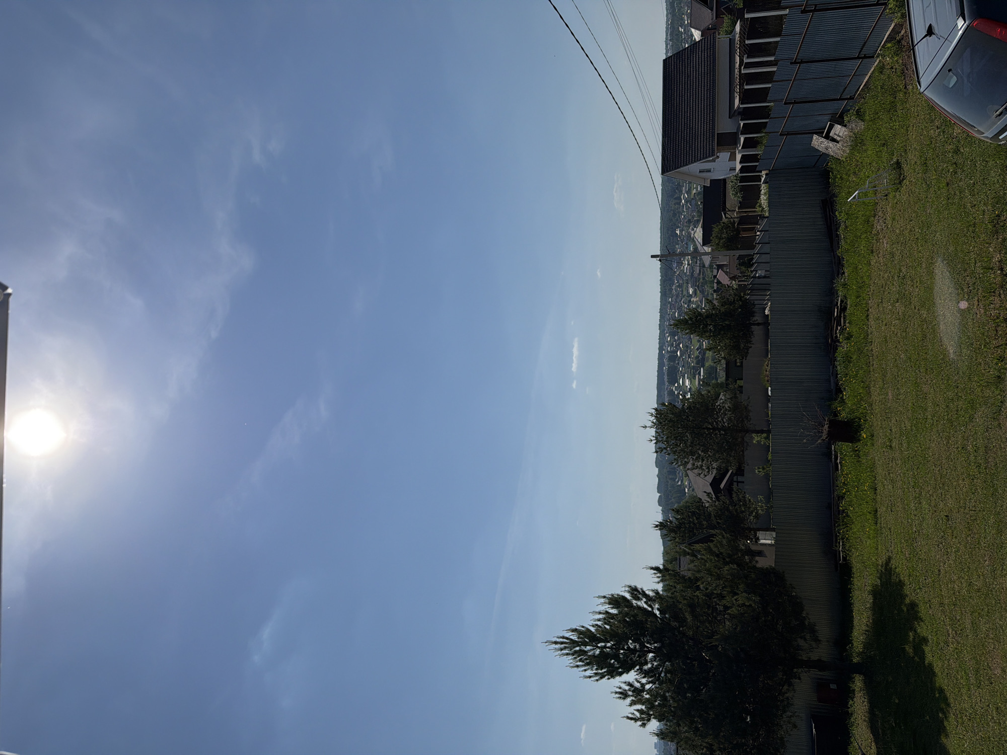 The photograph depicts a residential area on a sunny day. A house with a dark roof is visible, alongside a metal fence. Green grass covers the ground adjacent to the house. In the background, a cityscape stretches into the distance under a blue sky with scattered clouds. Utility poles and wires are visible above the houses. Based on location data, this scene is in Ягул, Russia. A blue car is parked to the right of the frame.