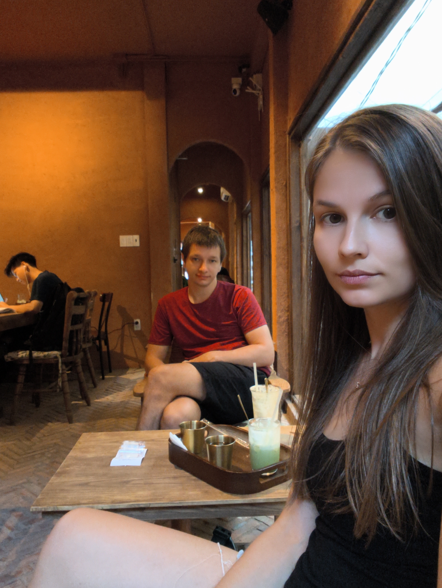 A selfie taken in a cafe shows a young woman in the foreground, facing the camera and looking directly into it. Her long, brown hair frames her face, and she is wearing a black tank top. To her left, a man in a red t-shirt and dark shorts is visible, looking slightly away from the camera. They are seated at a wooden table where two drinks, one appearing to be a layered matcha and milk beverage, and a tray with metal cups are placed. In the background, another man with earphones is working on a laptop at a table, with chairs and an arched doorway also visible. The cafe's interior features warm orange walls and wooden furniture, suggesting a cozy atmosphere. The scene is set in a cafe in Bac Nha Trang Ward, Vietnam.