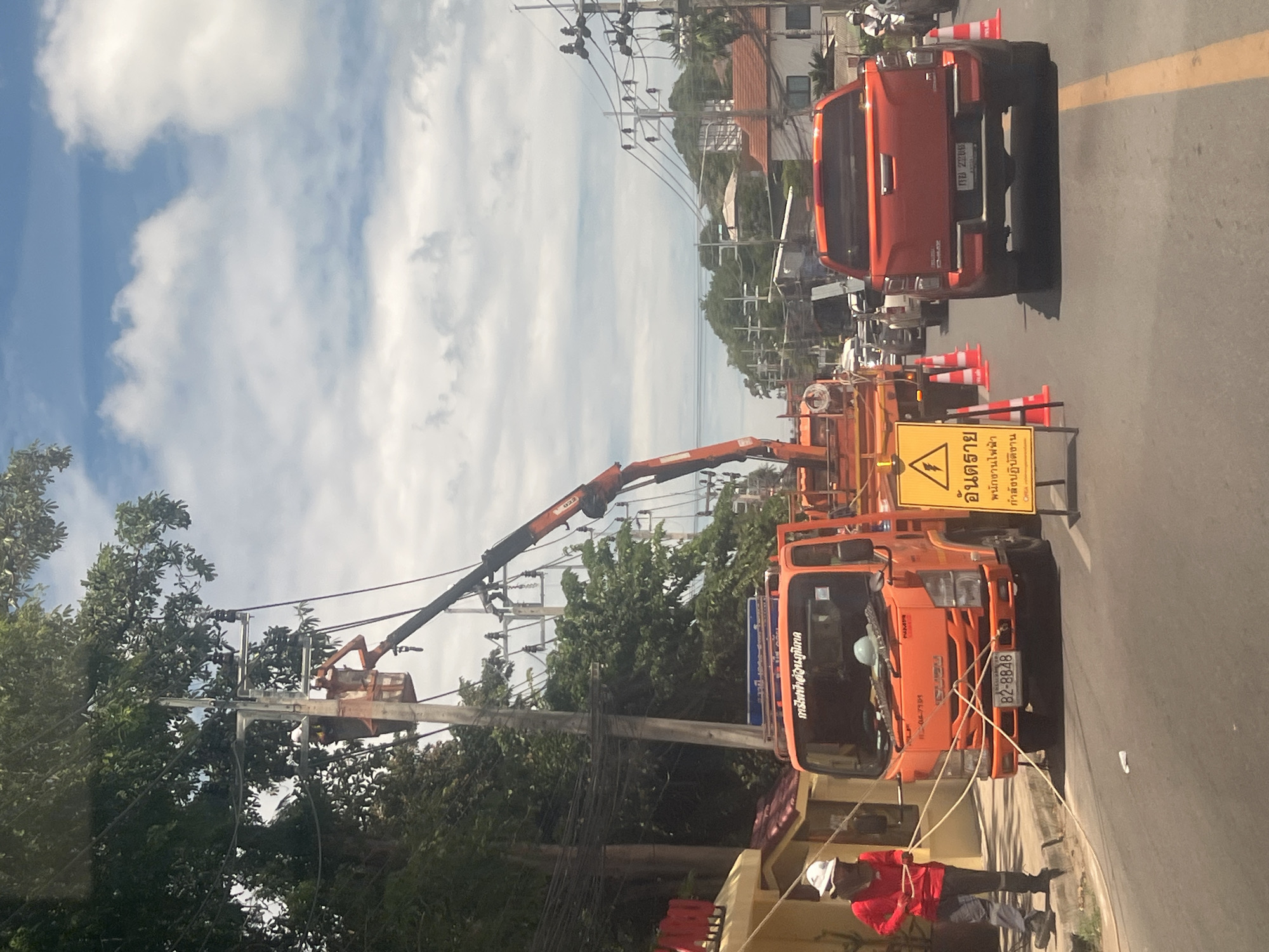In Ko Samui District, Thailand, on a public road, an orange utility truck with a crane is parked, with its bucket elevated to access overhead power lines. A worker in a red shirt and white hard hat is visible in the bucket, appearing to be performing maintenance. The truck has the license plate "82-8848" and displays Thai script. Adjacent to the truck is a yellow safety sign with a lightning bolt symbol, warning of electrical hazards and indicating work in progress. Orange traffic cones are placed around the work area. A red pickup truck is parked behind the utility truck, also showing Thai license plates. Another worker in a red shirt and white hard hat stands near the utility truck, holding a rope. The scene suggests ongoing electrical work on a street with trees, buildings, and traffic.
