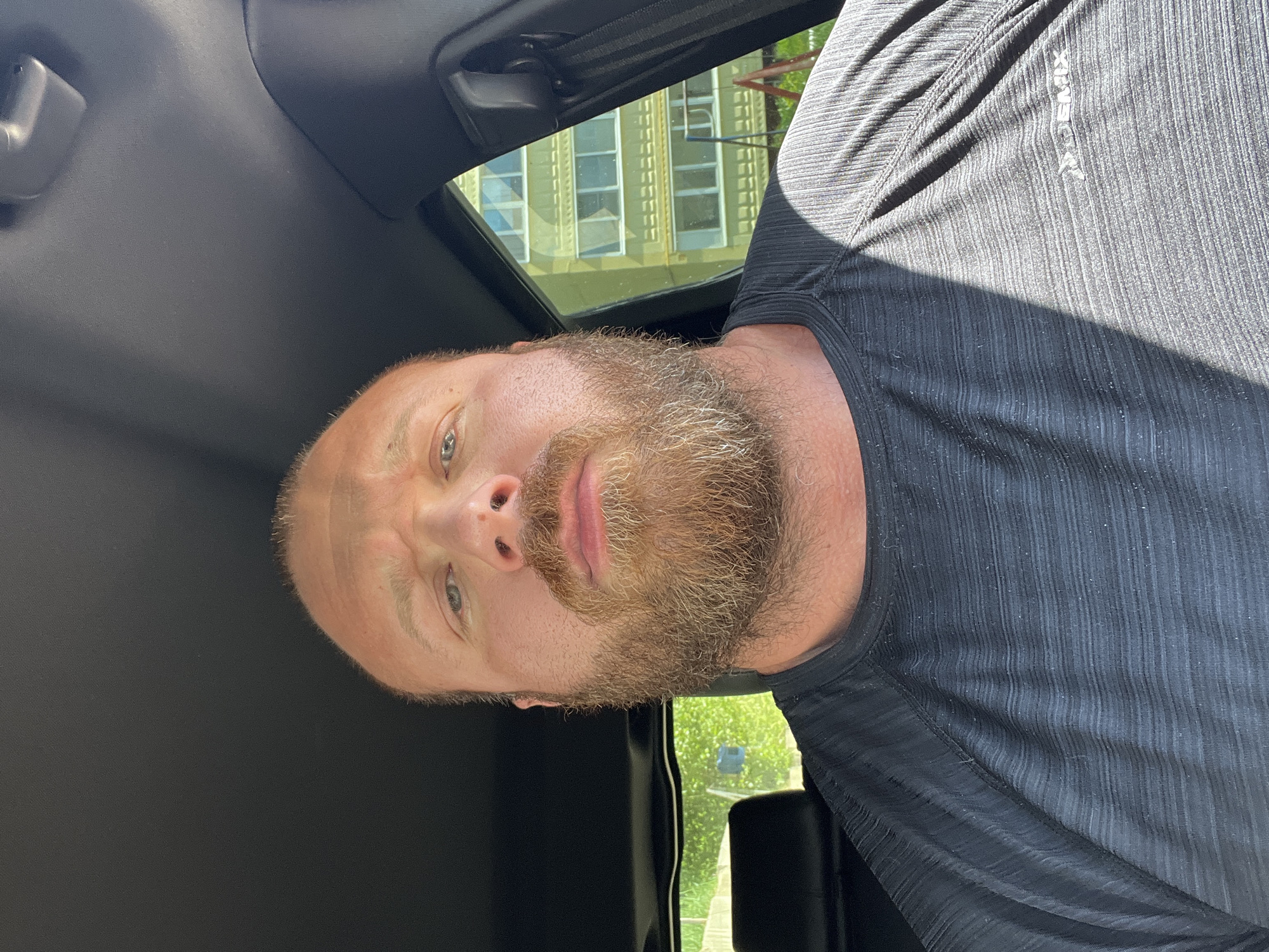 A man with a beard is in a vehicle, possibly a car, with an open sunroof. The man is wearing a dark-colored, possibly black, athletic shirt. Outside the vehicle, buildings with multiple windows are visible. Based on the image content and instructions, the location is assumed to be in the city of Noviy, Russia, though this cannot be confirmed with certainty from the visual information alone.