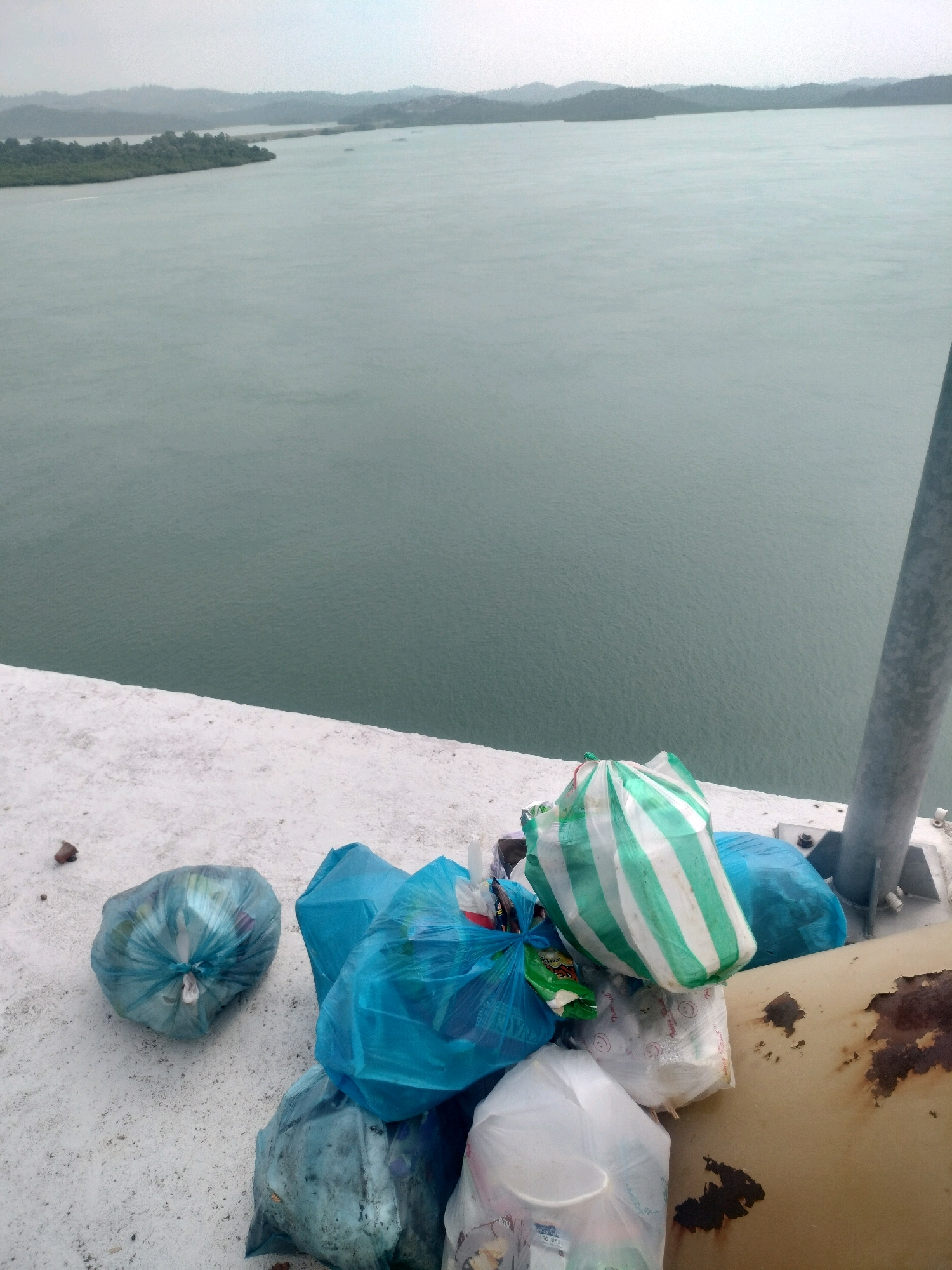 The image captures a scene on a white concrete surface overlooking a body of water. A pile of trash bags, some clear and others opaque blue and green with white stripes, are gathered near a rusty metal structure and a grey pole. The trash includes various items wrapped in plastic. In the background, a hazy landscape of islands with lush green vegetation and distant hills is visible across the tranquil, gray-green water. The lighting suggests an overcast day. This location is likely near the coastline of Batam, Indonesia, given the presence of islands and the tropical vegetation.