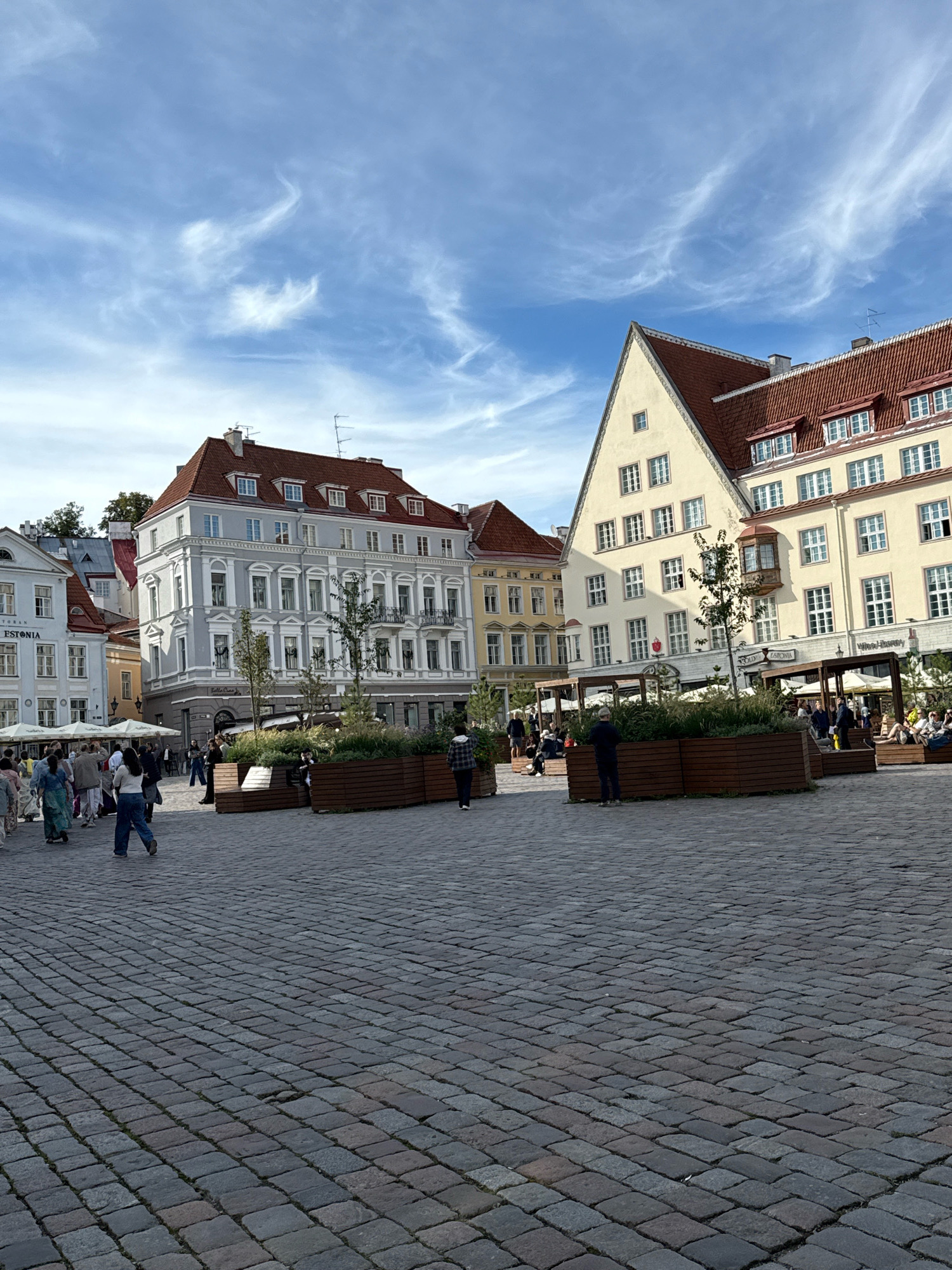 The image captures an outdoor public square paved with cobblestones. Several historic European-style buildings with red tiled roofs line the perimeter of the square. In the foreground, a woman in a blue shirt and dark pants is walking away from the camera, her legs and back visible. To her left, a group of people are walking and standing. In the middle ground, there are wooden planters with greenery and trees, and more people are seated or standing around them. In the background, the buildings feature multiple windows and architectural details. The sky above is blue with wispy white clouds. The scene appears to be a bustling urban environment on a clear day.