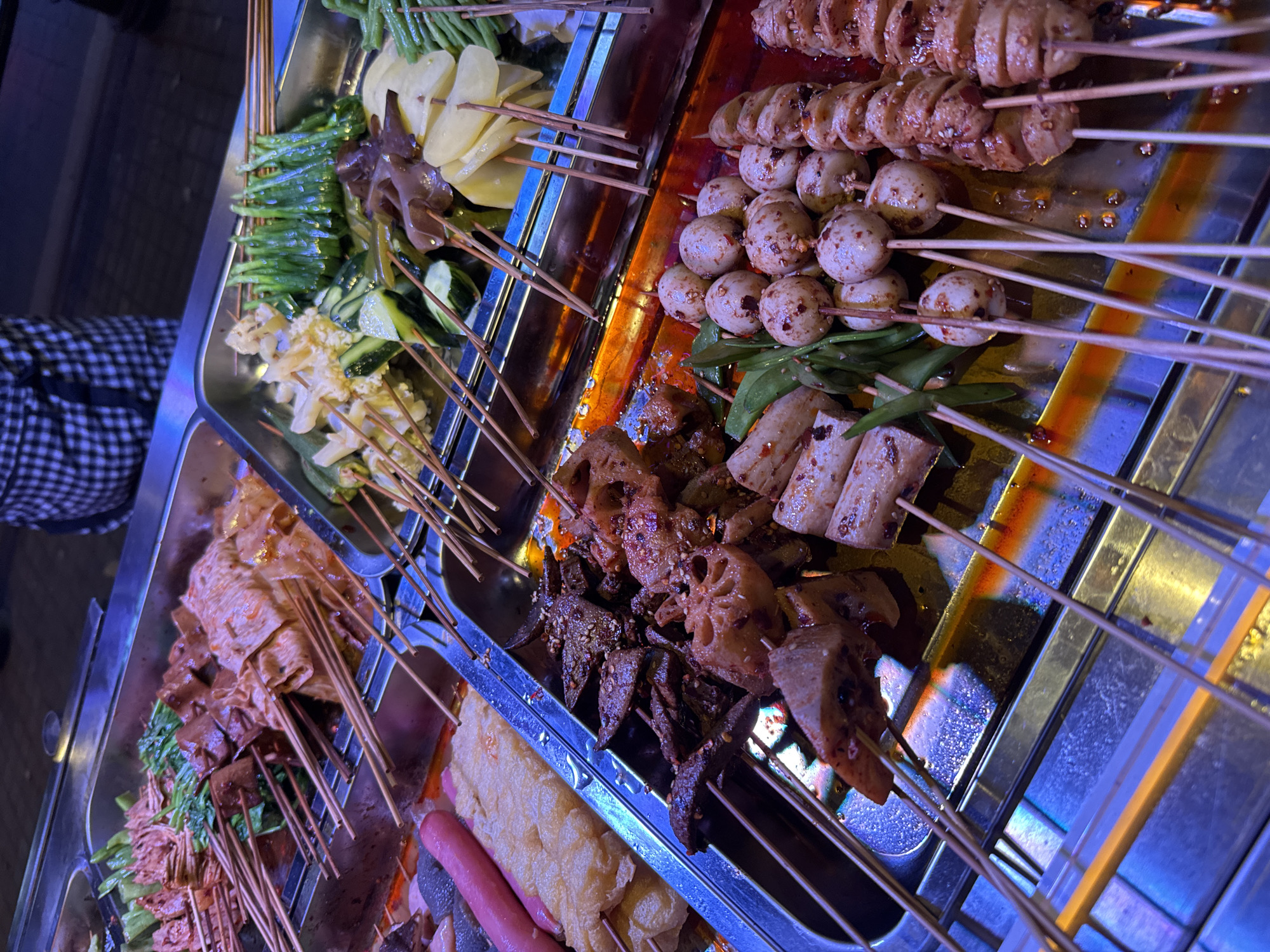 The image depicts a street food stall in Yuzhong District, China. Multiple stainless steel trays hold various foods on skewers. The first tray contains green beans, sliced potatoes, and mushrooms. Another tray has sliced sausage and meat balls. The third tray contains tofu, vegetables, and meat skewers. The skewers have a reddish sauce or seasoning. The food stall is illuminated by a blueish hue. The photo is taken at an angle, offering a partial view of the street and the person in plaid clothing to the left.