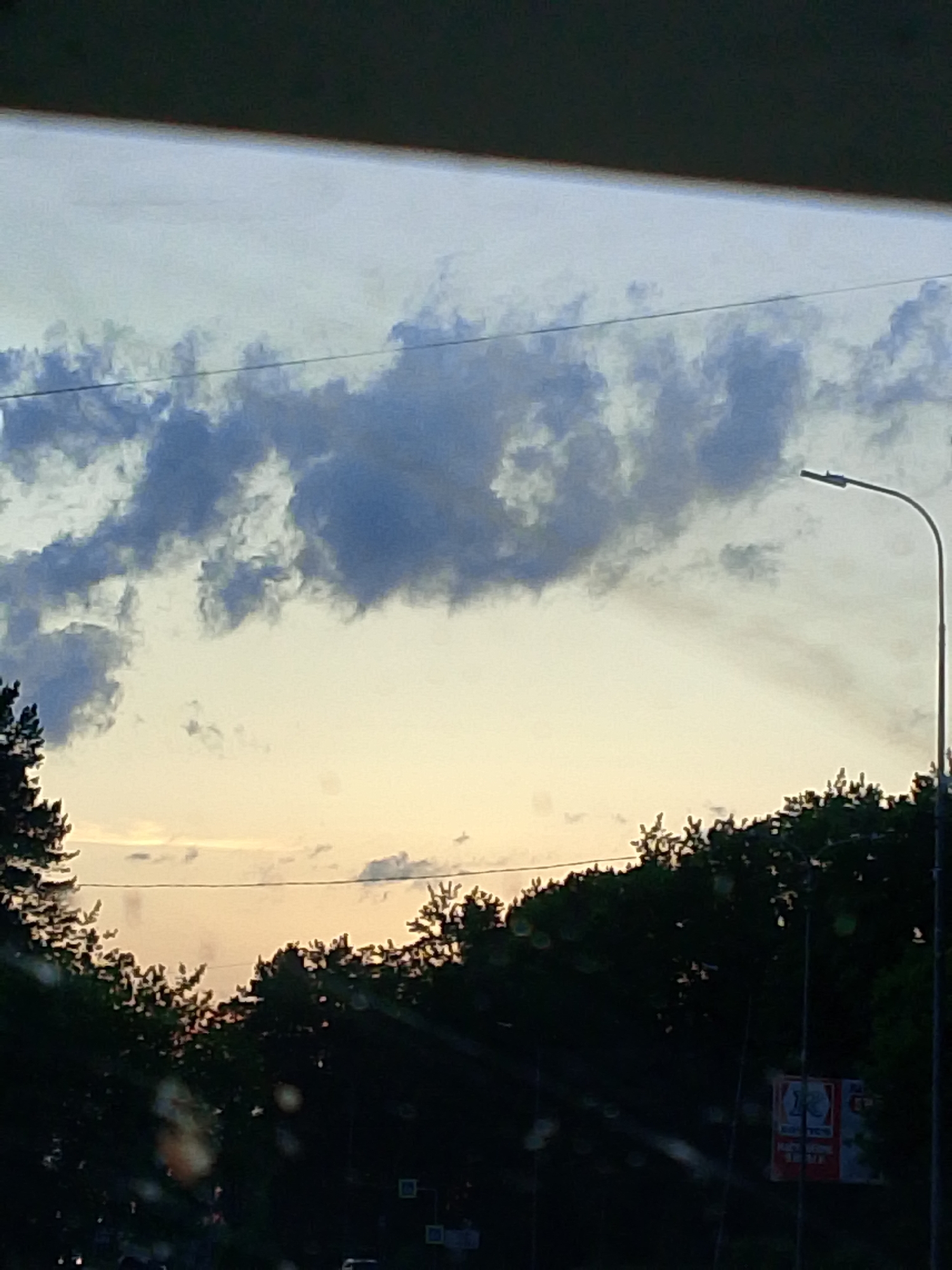 The image shows a view of the sky with clouds, partially obscured by an overhanging structure at the top of the frame. Below the sky, a line of trees is visible. A street lamp stands on the right side of the frame. In the lower portion of the image, there is a street with various signs and potentially traffic lights, though details are obscured. The photo appears to have been taken through a window, as there are water droplets visible in the foreground. Based on available information and the presence of distinctive signs, the location is in the city of Chaikovsky, Russia.