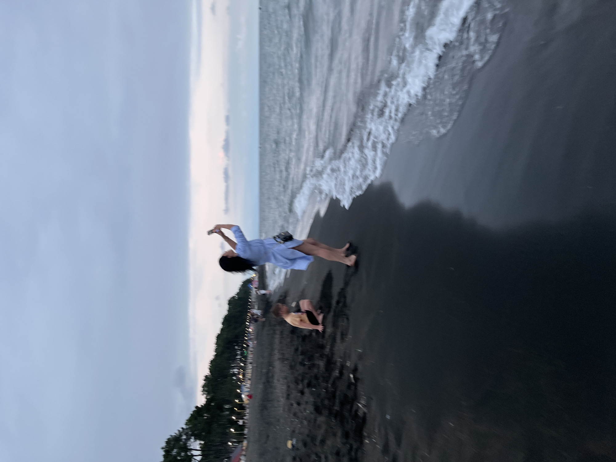 A woman in a light blue dress stands on a dark sand beach in Ureki, Georgia, holding a cell phone to take a picture. She is looking upwards, her body angled towards the sea. To her left, a young boy in a yellow t-shirt and black shorts is crouched down on the sand, also looking away from the camera. The sea is calm with gentle waves breaking on the shore. In the background, a line of trees separates the beach from a faintly lit area with what appear to be amusement park lights. The sky is overcast with hues of grey and pale pink. The overall impression is a serene evening at the seaside resort town.