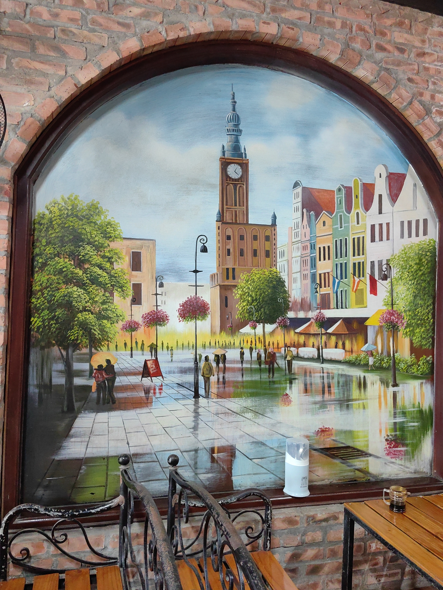 A detailed oil painting depicting a European city square is framed by an arched opening in a brick wall. The painting shows a tall clock tower at the square's center, surrounded by colorful buildings with peaked roofs, reminiscent of a European architectural style. It appears to be a rainy day, as the pavement is wet and reflective. Several figures are visible in the painting, some walking with umbrellas. Street lamps adorned with flower baskets line the square.
In the foreground, part of an ornate iron chair and a wooden table are visible, suggesting a cafe setting. A utensil holder and a small glass mug sit on the ledge beneath the painting. The brick wall surrounding the painting suggests an interior setting with a rustic aesthetic. There is no concrete evidence to confirm the location of the image in Hà Nội, Vietnam.