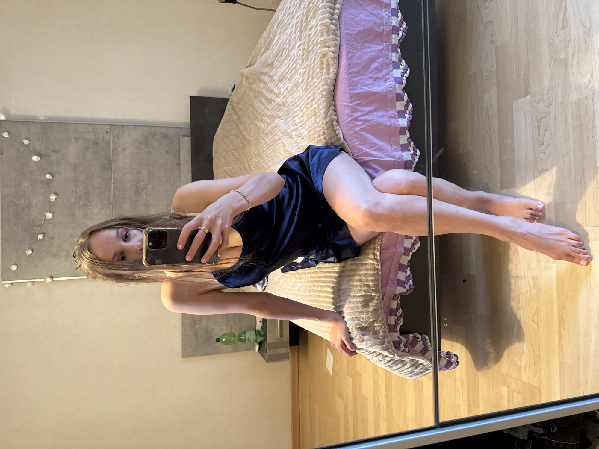A young woman with blonde hair tied in a ponytail, wearing a dark blue satin slip dress and a gold ring, is taking a mirror selfie in a bedroom. She is sitting on the edge of a bed covered with a beige textured blanket and a pink fitted sheet with a white and purple border. To her left, a nightstand with a grey top holds a green bottle. The room has light-colored walls and wooden flooring. Decorative lights are strung across the wall behind her. The mirror in the foreground reflects her lower body and feet, along with a shadow. The scene appears to be in a private residence.