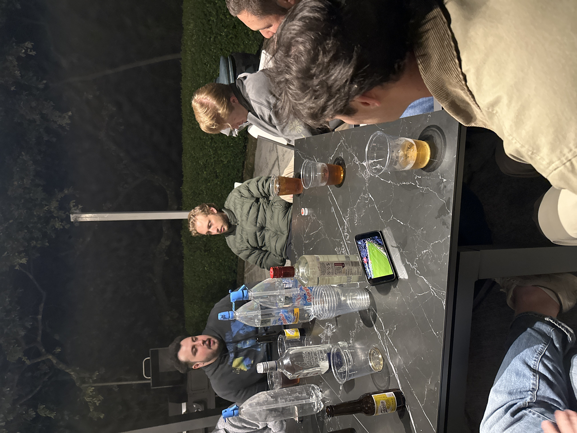 The image depicts a group of five men gathered around a black marble-patterned table outdoors at night. The table is cluttered with empty plastic bottles, liquor bottles, plastic cups containing beer, and a smartphone displaying a soccer match. The men are looking at the phone and the contents on the table. One man wears a green jacket, another a gray hoodie, and another a tan jacket. The location, city of Mexico City, Mexico, is not discernible from the given image.