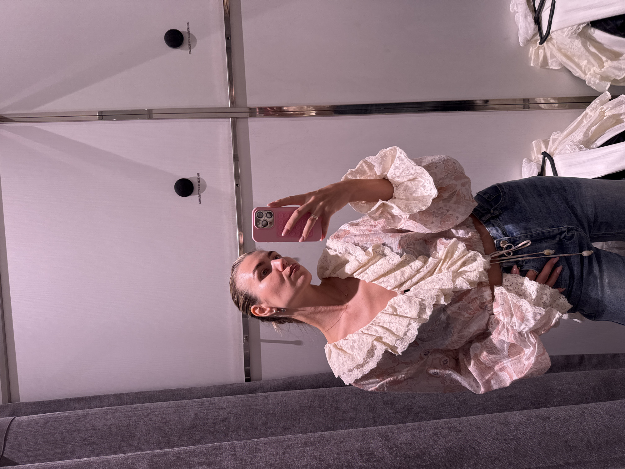 A woman is taking a selfie in front of a bank of white lockers. She is wearing a ruffled lace top, jeans, and holding a pink phone. The environment appears to be indoors, with a gray surface below the lockers.