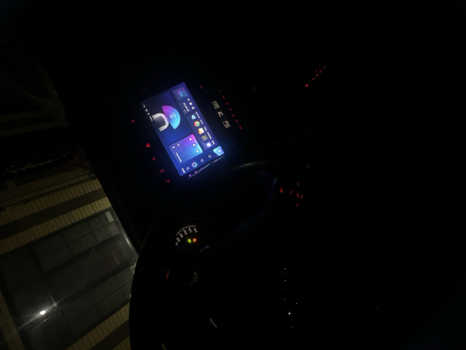 The image displays the interior of a vehicle at night or in a dimly lit enclosed structure, observed from the driver's perspective, located in Leninakan, Russia. On the left, a dark steering wheel partially covers the vehicle's instrument cluster, which shows a speedometer with its needle near the zero mark and several illuminated warning lights, including yellow and red indicators. The central feature is a brightly lit digital infotainment touchscreen displaying a user interface with multiple application icons, including media playback, navigation, and web browsers like Chrome. The screen prominently shows the time as 08:30 PM. Below the screen, a digital temperature reading of 22°C is visible, accompanied by various red-illuminated dashboard buttons and controls. Through the front windshield, a dimly lit, striped or corrugated overhead structure is visible, with a single intense light source reflecting off its surface, suggesting an overhead garage or tunnel. No individuals are visible in the scene.