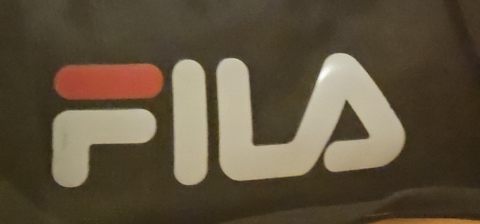 The image displays the Fila logo prominently. "Fila" is spelled out in a bold, sans-serif font. The "F" is unique, featuring a white lower section and a red horizontal bar at the top. The letters "I", "L", and "A" are all white. The logo has a slightly raised, glossy appearance, and it is set against a dark gray background. The letters look inflated and rounded at the ends.
