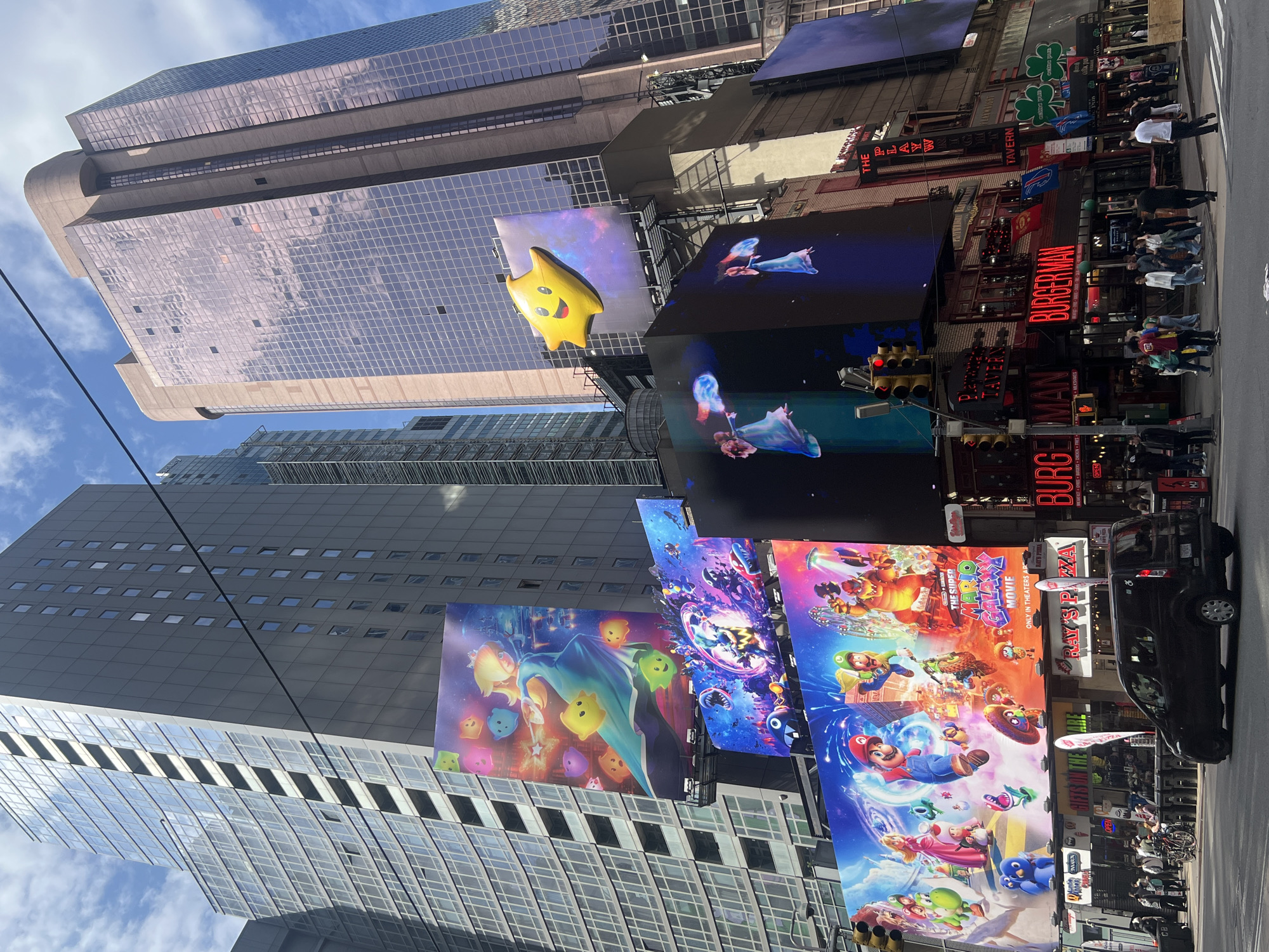 The image shows a busy street scene in Times Square, New York City, United States of America. Several towering buildings are visible, including one with a reflective glass facade and another with a grid-like window pattern. Large electronic billboards display images from "The Super Mario Galaxy Movie" and other advertisements, including a BurgerMan restaurant advertisement. A black vehicle is parked along the side of the street. Pedestrians are seen walking along the sidewalks, and a few are crossing the street. Restaurant signage, including Ray's Pizza and the Playwright Tavern, are also visible. The sky is partly cloudy.