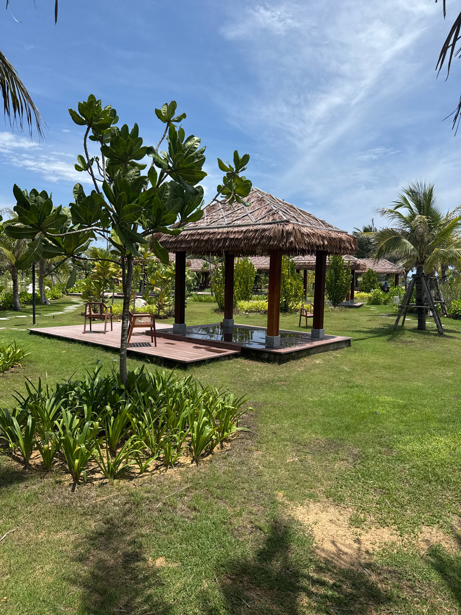 A thatched-roof gazebo with wooden pillars and a water feature is set in a well-manicured lawn. Two wooden chairs are placed on a wooden deck in front of the gazebo. Palm trees and tropical foliage are visible in the background, with a clear blue sky overhead. This scene is located in the coastal city of Cam Ranh, Vietnam.