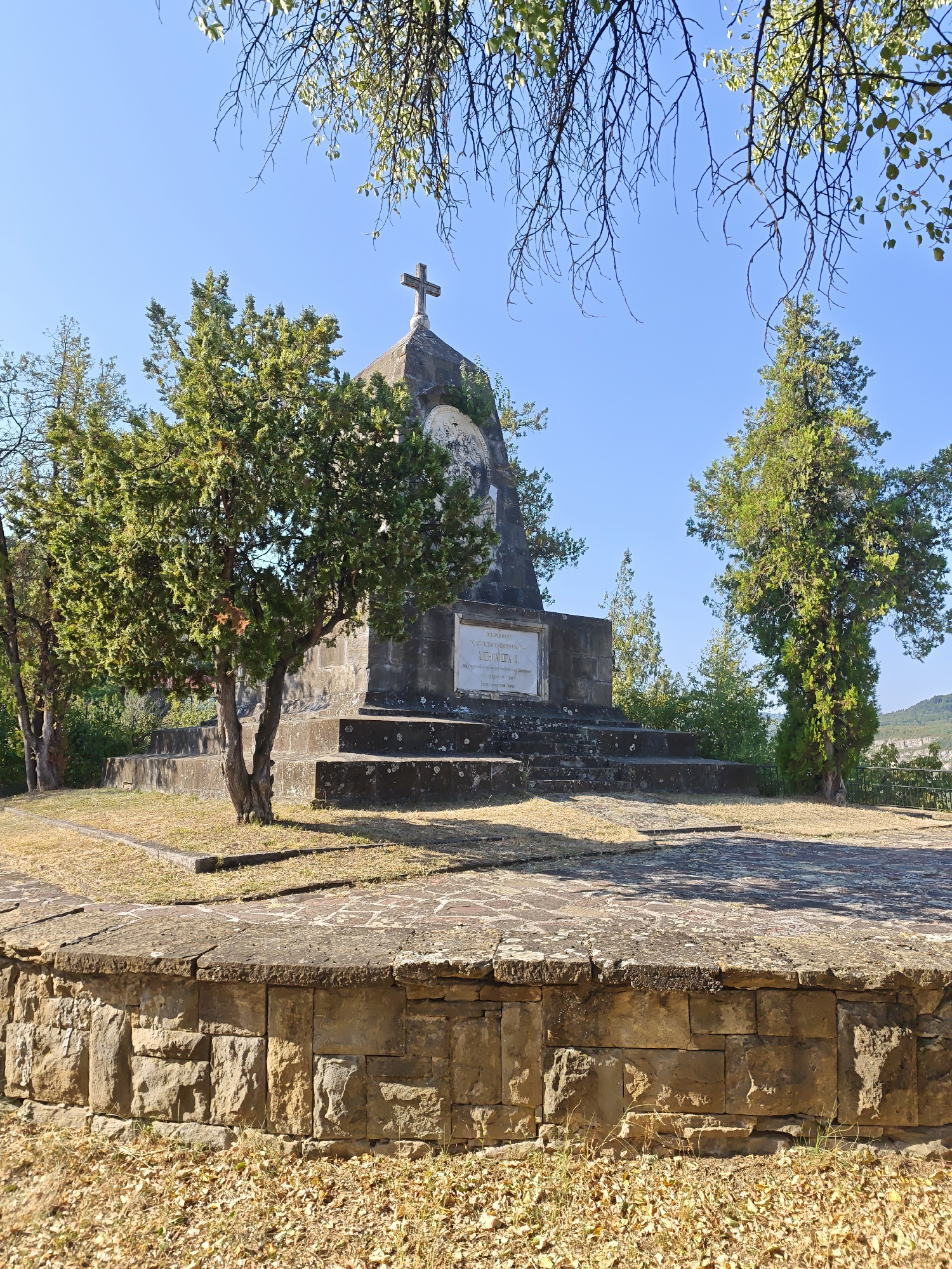 This image shows a monument with a cross on top, set against a clear blue sky. The monument is made of stone and has a circular base with steps leading up to it. There are trees and dry grass around the monument. In the background, there are hills and more trees. The monument is located in the city of Lovech, Bulgaria, and is dedicated to Tsar Ivan Shishman. It stands on a hill overlooking the city.