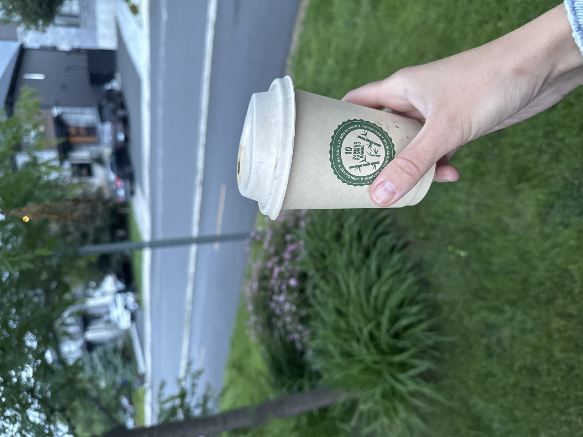 A close-up shot shows a hand holding a disposable coffee cup made of a light brown, biodegradable material. The cup has a white lid and features a green circular logo with text and an illustration of bamboo stalks. The background is blurred, but it appears to be an outdoor setting with a street, cars, and houses visible. The overall impression is a casual moment, possibly in a residential area of Gatineau, Canada, given the architectural style of the houses and the presence of mature trees and manicured lawns. The lighting suggests it is either early morning or late afternoon.