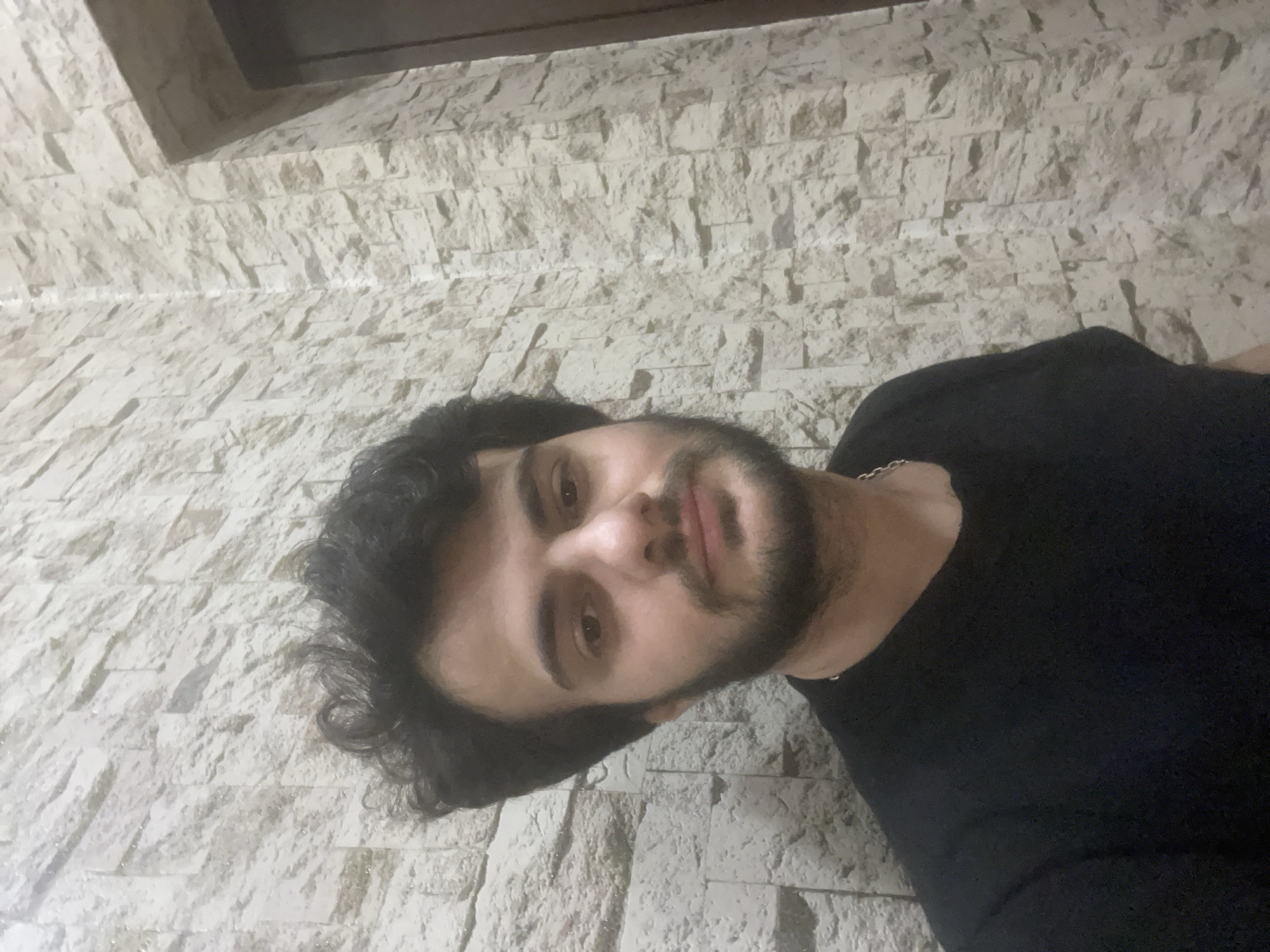 A medium shot shows a young man with dark, curly hair and a beard standing in front of a textured, light-colored stone wall. He is wearing a black t-shirt and a silver chain. The background also shows a portion of a dark door frame on the right side. The scene does not contain any information that would indicate its location in Tbilisi, Georgia.