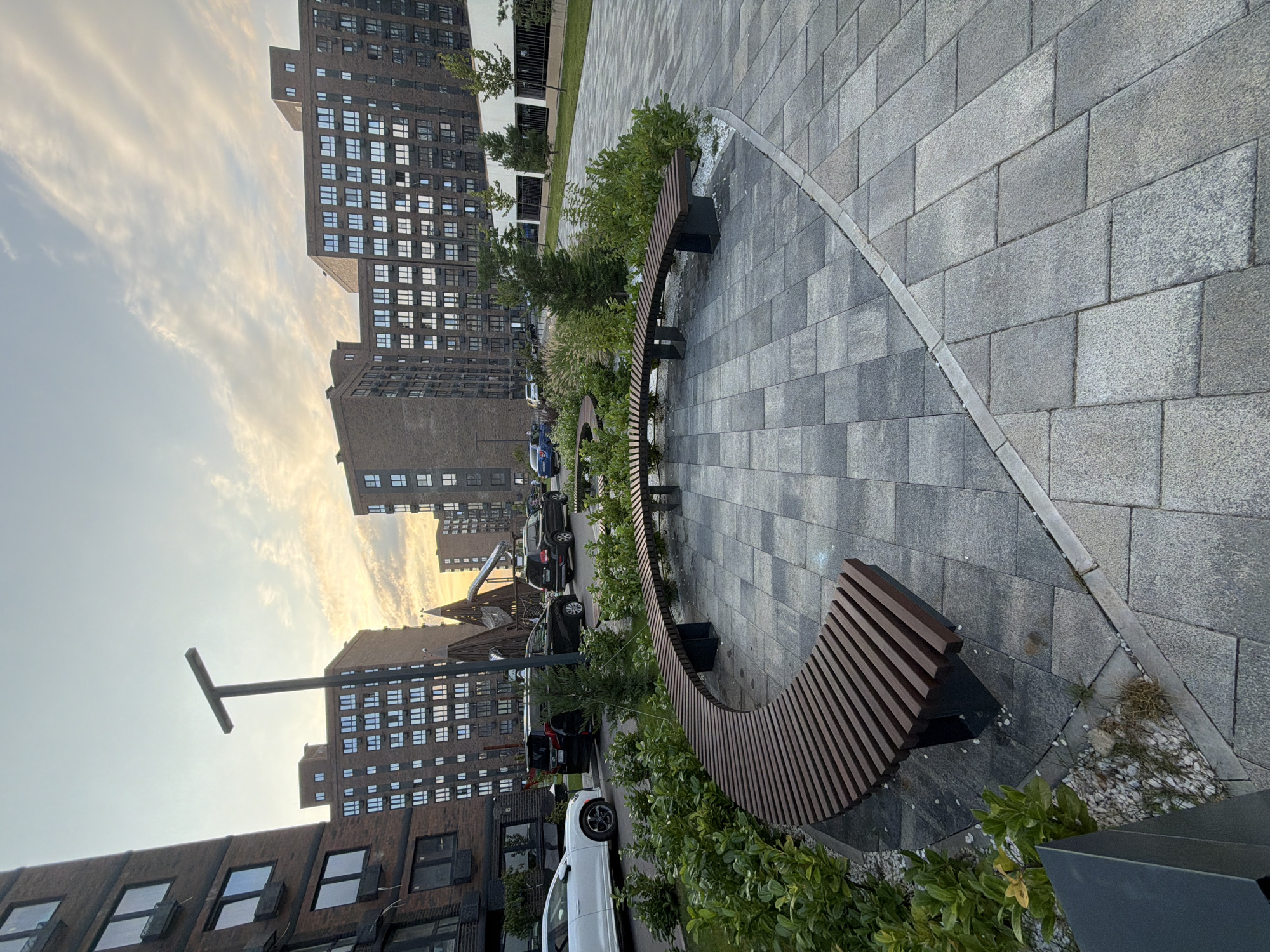 In a city courtyard, a curved wooden bench is the focal point, set on a paved area with grey rectangular tiles. To the left of the bench, lush green bushes and small white stones create a decorative border. In the background, several modern apartment buildings with numerous windows are visible, under a sky with scattered clouds at dusk. To the right, a pathway leads towards a white building with a fence. Several parked cars, including a white sedan and black SUVs, are lined up on a street adjacent to the courtyard. A lamppost with a T-shaped head stands to the left of the bench.