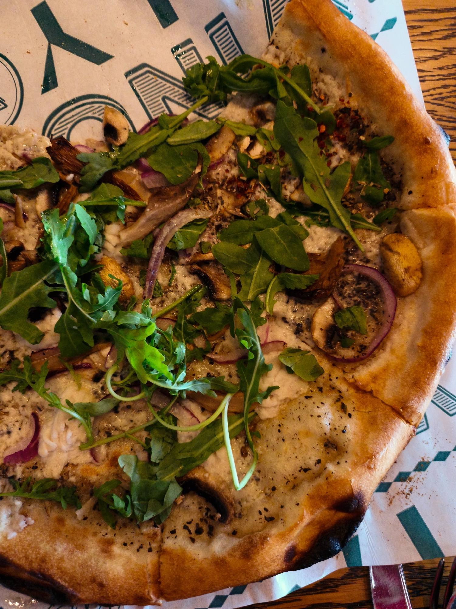 A close-up, overhead shot captures a partially eaten pizza placed on a patterned paper liner on a wooden table. The pizza features a golden-brown crust and is topped with melted cheese, fresh arugula, mushrooms, and thinly sliced red onions. Scattered red pepper flakes and dried herbs are visible across the surface. The background shows a portion of the paper liner with a stylized green geometric design, and a wooden surface. The scene is set in Be'er Sheva, Israel.