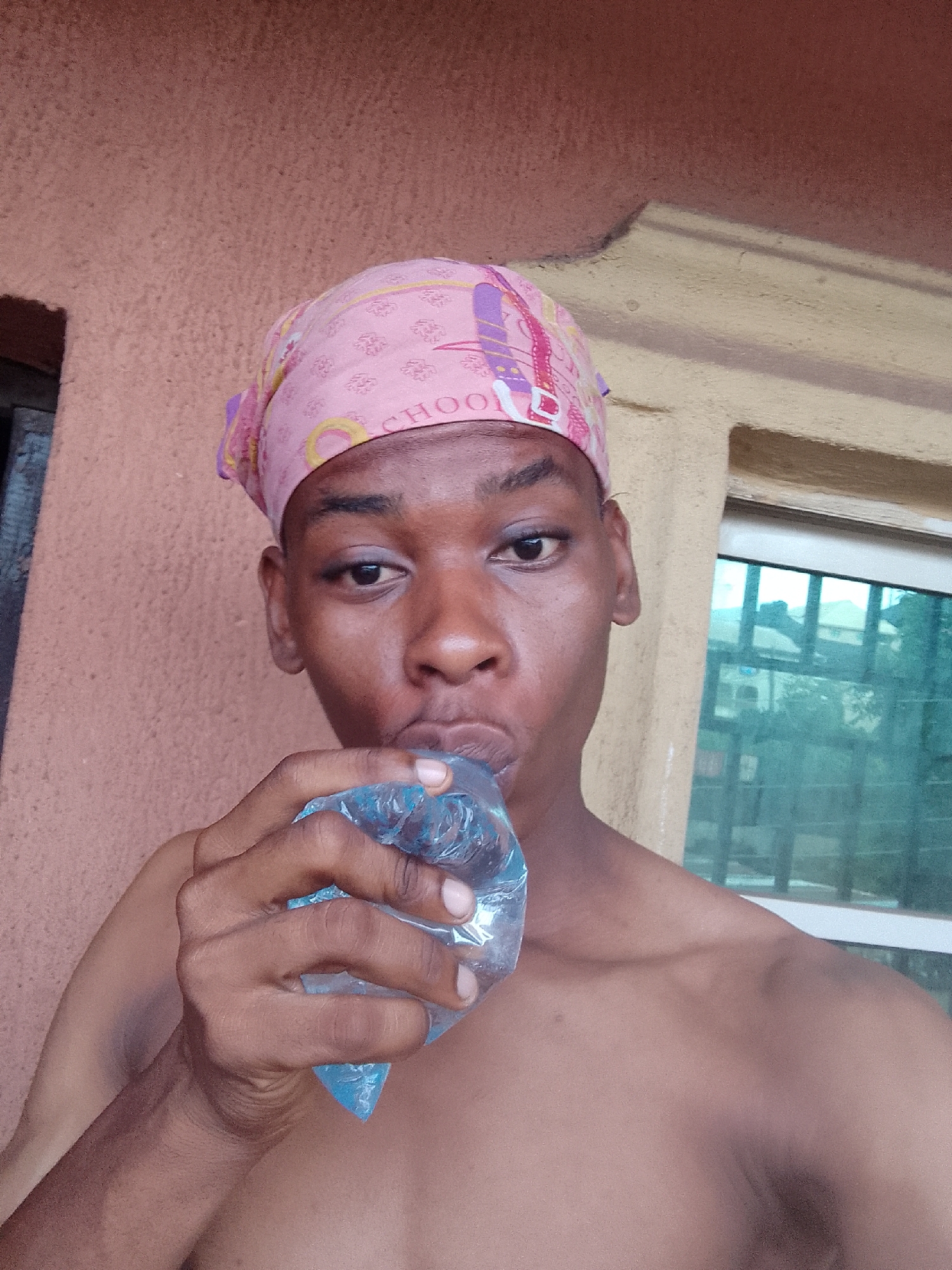 A shirtless young man wearing a pink headscarf with cartoon character designs is shown from the chest up, holding a small, sealed, clear plastic bag filled with blue liquid to his mouth as if he is drinking from it. He is facing the camera with a serious expression. Behind him is a peach-colored wall on the left and a window on the right showing the reflections of outdoor buildings and trees. The location is the city of Unknown, Unknown.