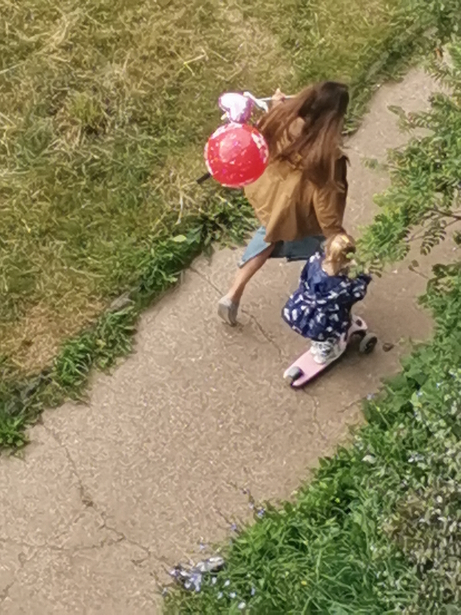 An adult with long brown hair, wearing a brown jacket and blue shorts, walks along a concrete path, holding a bunch of balloons, one of which is red and round, and two are pink and heart-shaped. Beside the adult, a young child with blonde hair is riding a pink scooter. The child wears a dark blue dress with a white pattern. The path is bordered by green grass on one side and dense green foliage on the other. The scene appears to be outdoors, likely in a residential area or park, possibly in Chaikovsky, Russia, given the context of previous images. The lighting suggests it is daytime.