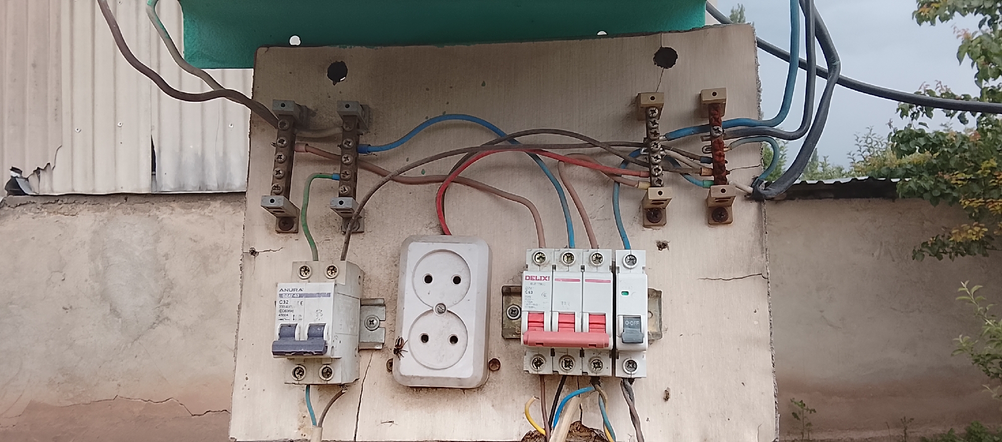 An outdoor electrical box is mounted on a weathered wooden board attached to a concrete wall. The box contains two circuit breakers, a double electrical outlet, and several terminal blocks for wiring. Wires of various colors, including blue, red, green, and brown, are connected to these components. A small spider is visible on the electrical outlet. The background shows a corrugated metal structure, a concrete wall, and foliage from trees. The sky is overcast.