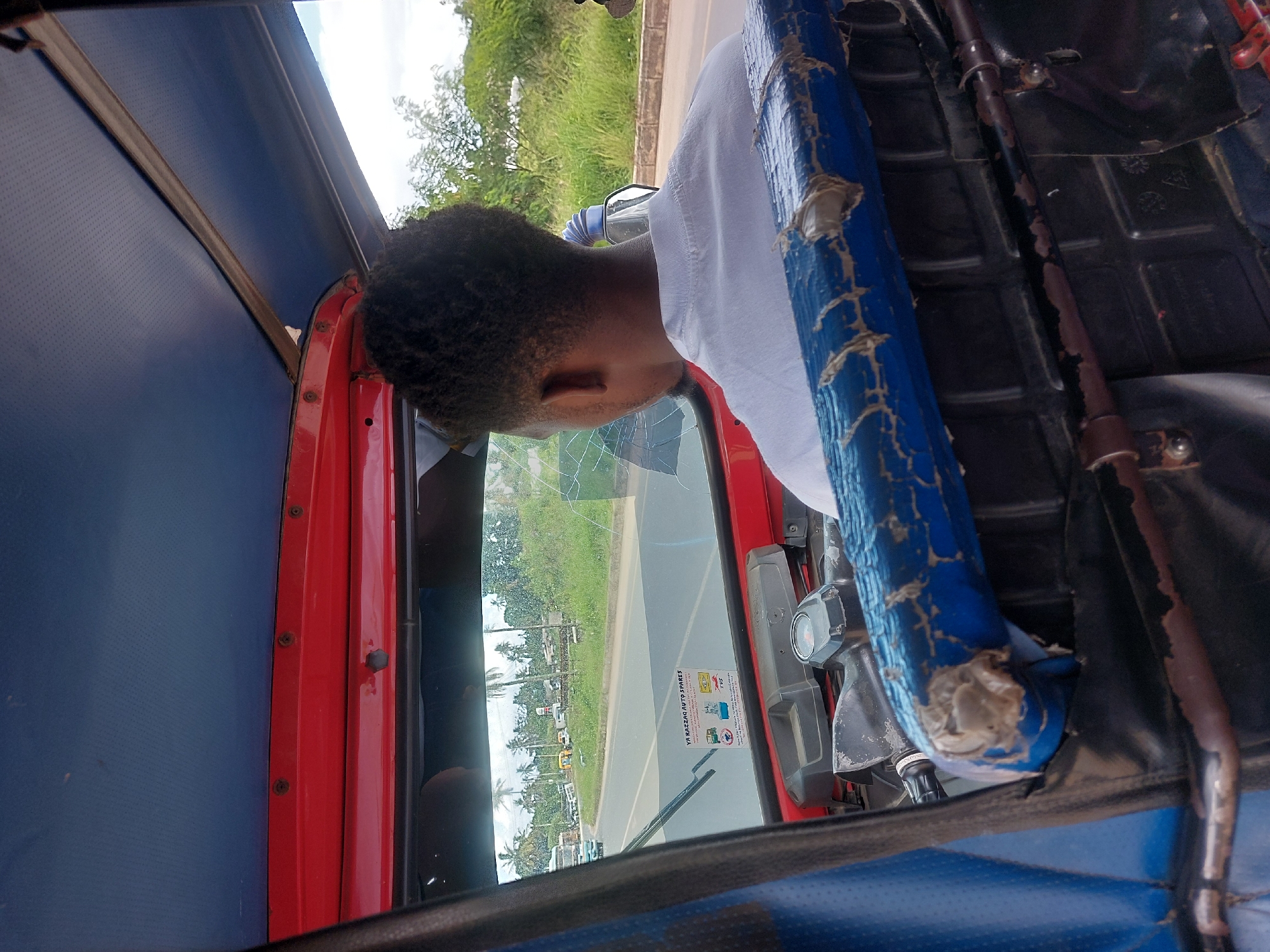 A person wearing a white t-shirt is driving a red and blue bajaj (a three-wheeled motorized vehicle) down a road in Dar es-Salaam, Tanzania. The bajaj has a cracked windshield. In the background, there are palm trees and other vegetation, as well as other vehicles.