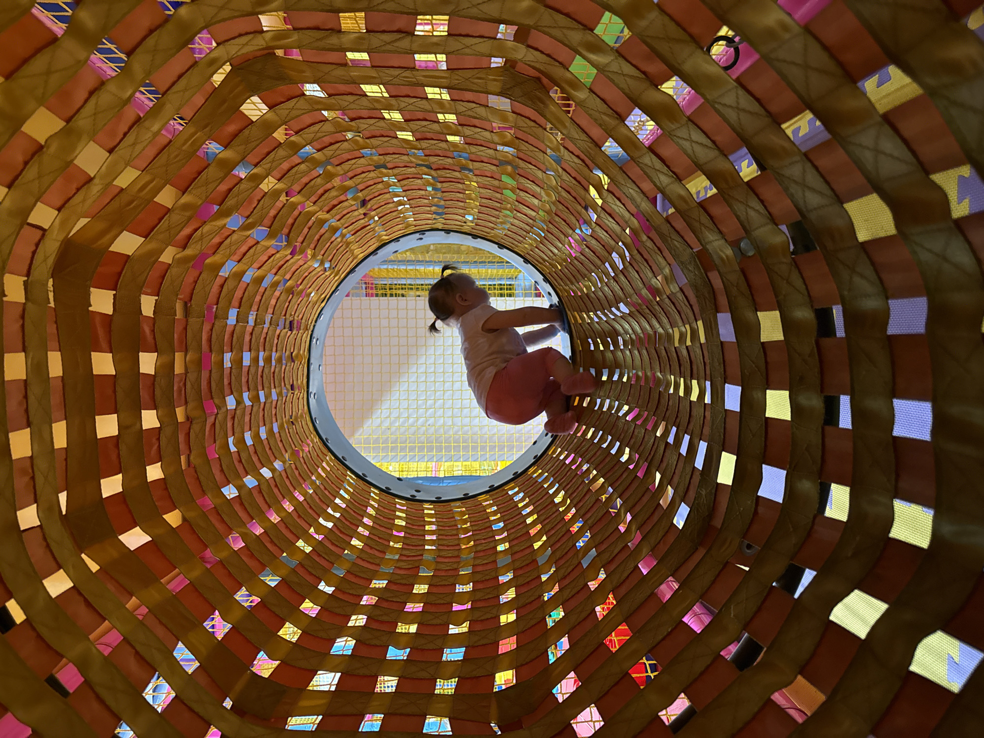 A child with a ponytail, wearing pink clothing, is navigating through a tunnel-like structure at an indoor playground. The tunnel is constructed with interwoven straps, creating square openings that allow light to pass through. The scene is illuminated with natural light filtering through the openings in the tunnel. The setting appears to be an indoor play area. The location is Kommunarka, Russia.