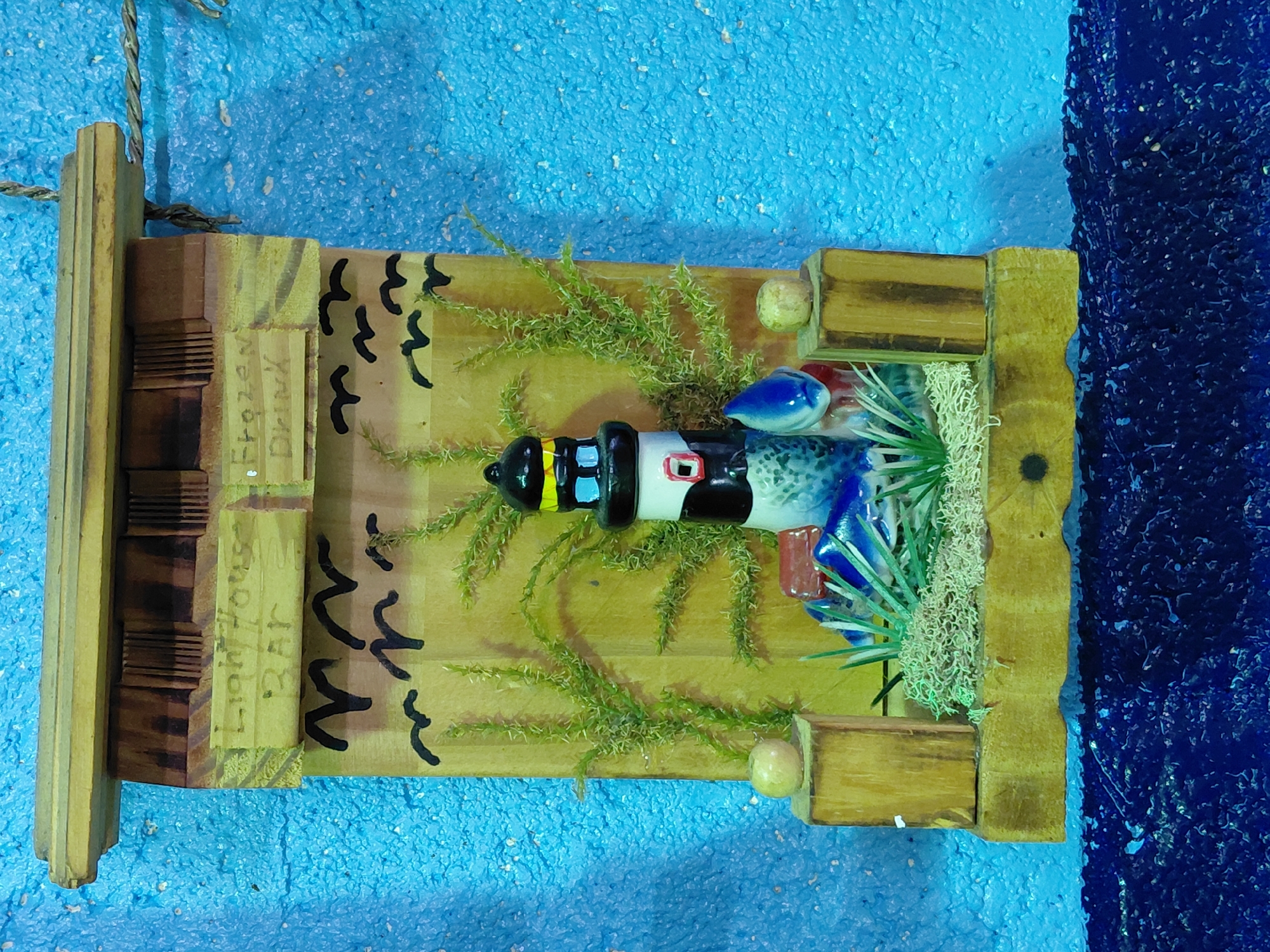 A rustic, wooden wall decoration, likely found in Cutler Bay, United States of America, depicts a stylized lighthouse scene. The piece is constructed from various wooden scraps, arranged to form a small diorama. A ceramic lighthouse model, painted with black, white, red, and blue sections, lies horizontally within a central opening, suggesting it might represent a fallen or capsized lighthouse. Patches of artificial moss and green, sprig-like foliage are scattered around the lighthouse, possibly intended to represent land or vegetation. Black painted "V" shapes, possibly meant to represent seagulls, are visible on the wooden backdrop. To the left of the lighthouse scene, a small wooden block is labeled "Lighthouse Bar Frozen Drink", indicating a theme of a seaside bar or establishment. The entire assemblage is mounted on a rectangular wooden base with a hanging rope at the top. The backdrop against which the piece is displayed is a blue, textured surface, with a darker blue section visible on the right.