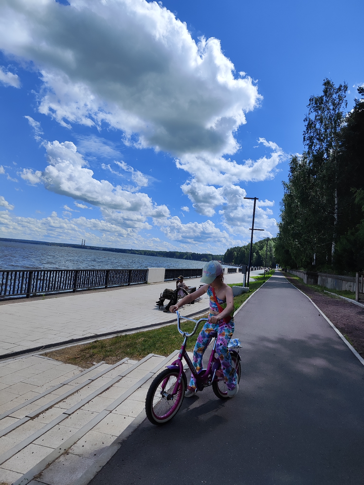 The image depicts a girl on a pink bicycle on a paved path next to a body of water. The girl is wearing a colorful patterned outfit and a baseball cap. The path runs alongside a railing and a sidewalk, which overlooks the water. Beyond the water, a treeline is visible, and in the distance, two smokestacks with condensation plumes are present. The sky is blue with scattered white clouds. The scene appears to be a park or promenade area. The presence of the smokestacks suggests an industrial or power generation facility nearby, which is consistent with the location of Chaikovsky, Russia, a city known for its hydroelectric power station. The image shows a sunny day with good visibility.