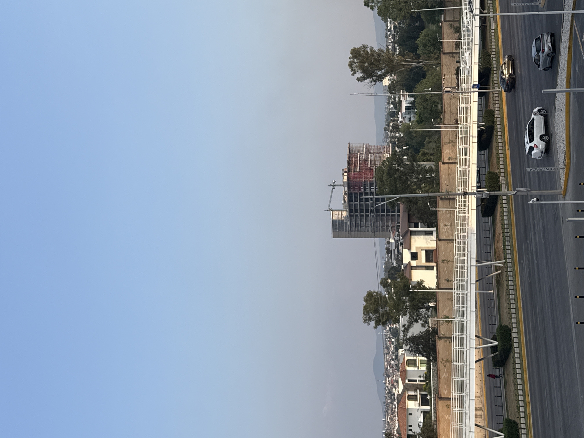 The image shows a view of San Bernardino Tlaxcalancingo, Mexico, with a multi-lane road running alongside a residential area and a large construction site in the distance. Several vehicles are visible on the road. The sky is clear and blue.