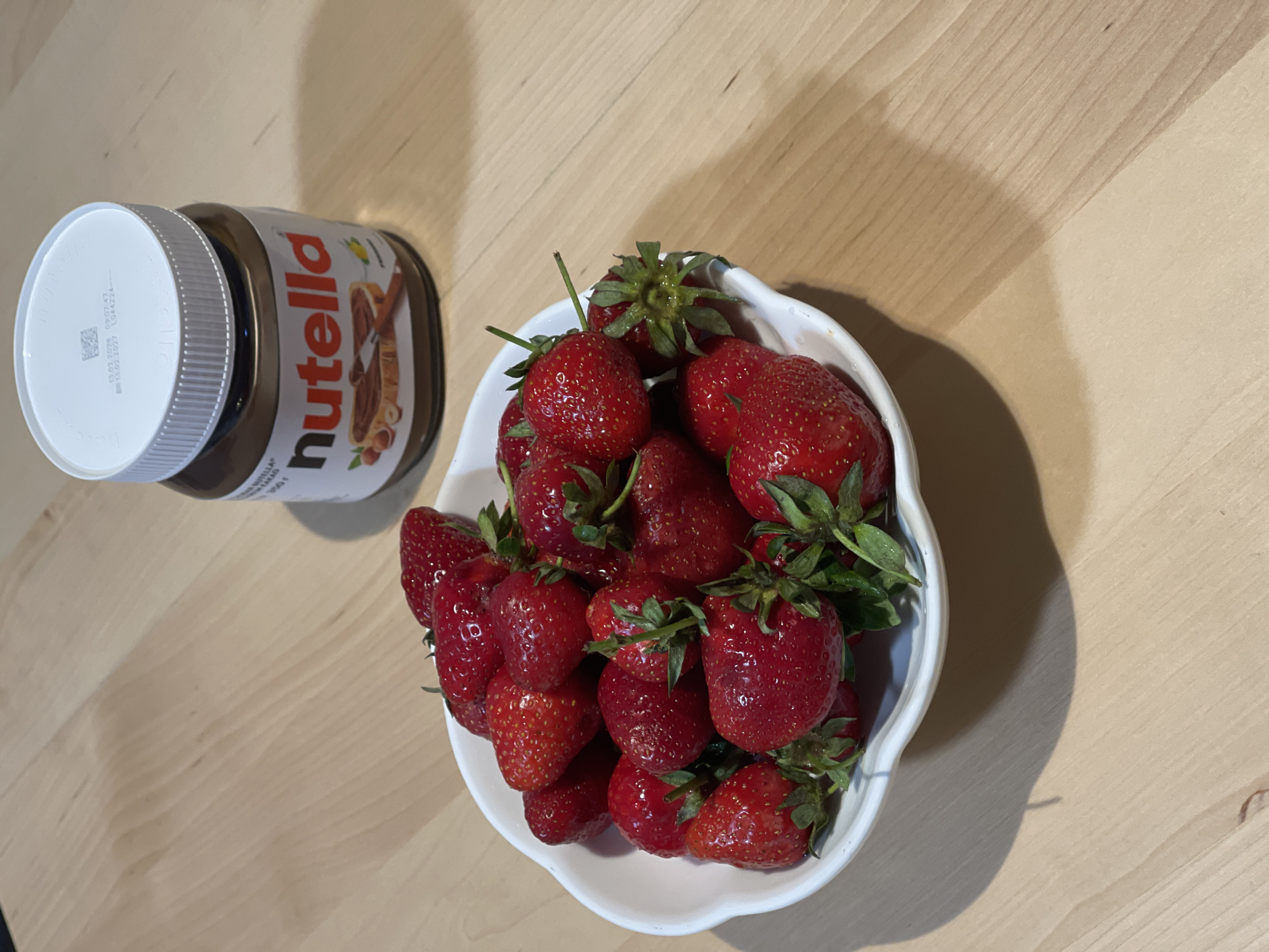 The image shows a jar of Nutella and a white bowl filled with strawberries on a light-colored wooden surface. The strawberries are red and have green stems. The Nutella jar is unopened. The setting appears to be indoors. There is no information available in the image to indicate its location as Izhevsk, Russia.
