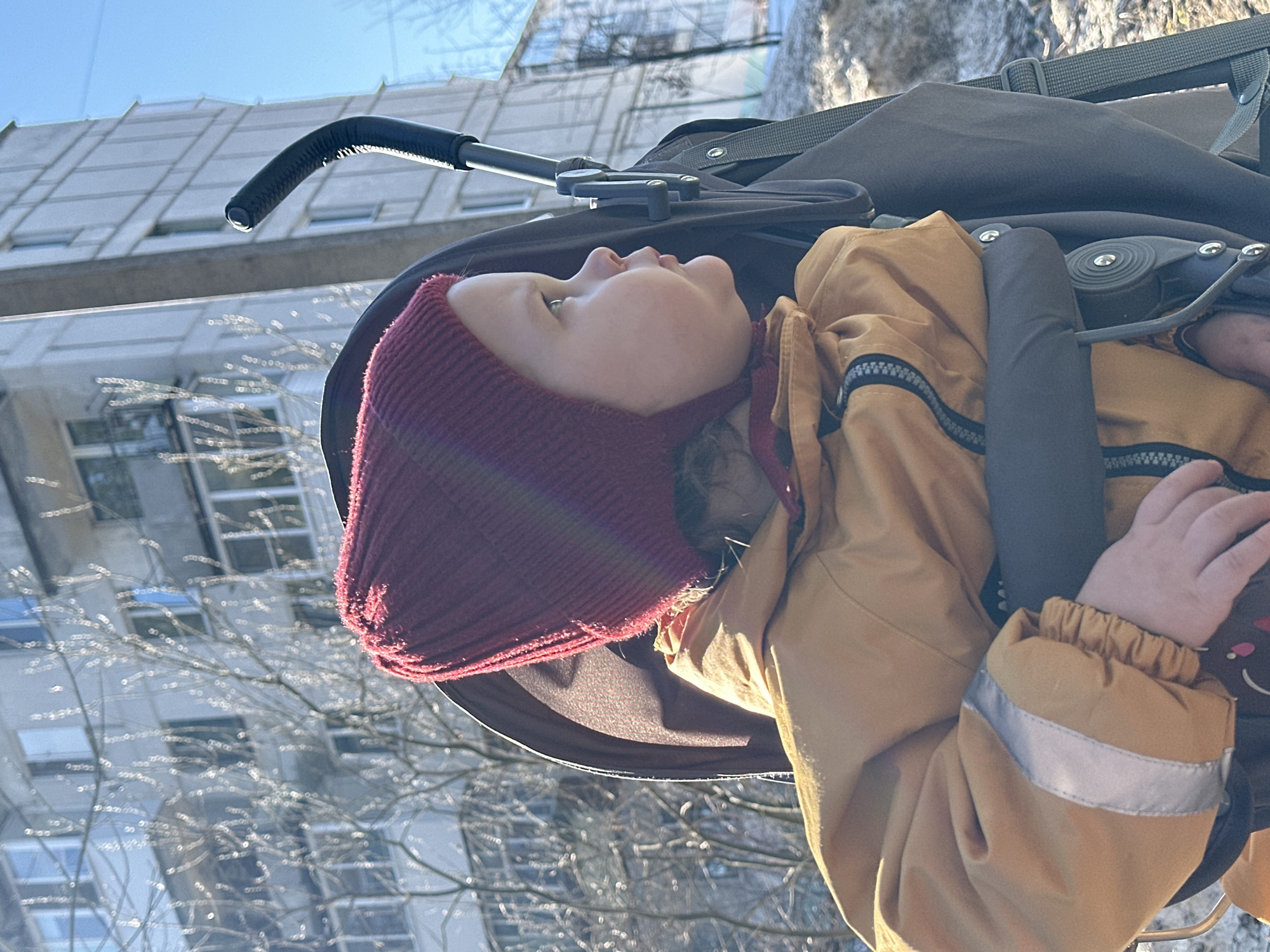 A child wearing a maroon knit cap and a tan jacket is sitting in a stroller. The stroller's handlebar is visible, and the child's face is turned upwards. The stroller is outdoors, with a building in the background that appears to be located in Chaikovsky, Russia, given the typical architecture. The lighting suggests it is daytime, and bare trees are also visible in the background.