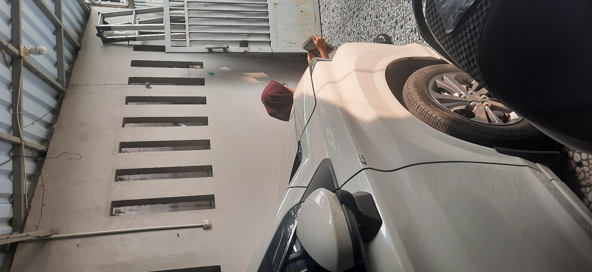 A white sedan is parked in an outdoor covered area with a corrugated metal roof supported by metal beams. The wall behind the car has vertical rectangular openings covered with what appears to be grating. A person wearing a maroon headscarf is leaning over the hood of the car, looking down at a tablet or notebook they are holding. To the right of the car, a silver metal gate is visible, and a ladder leans against the wall. The ground is paved with small, irregularly shaped stones. A portion of a black motorcycle is in the foreground. The lighting suggests it is daytime, with shadows cast by the roof and objects. The location is identified as Cisauk, Indonesia.