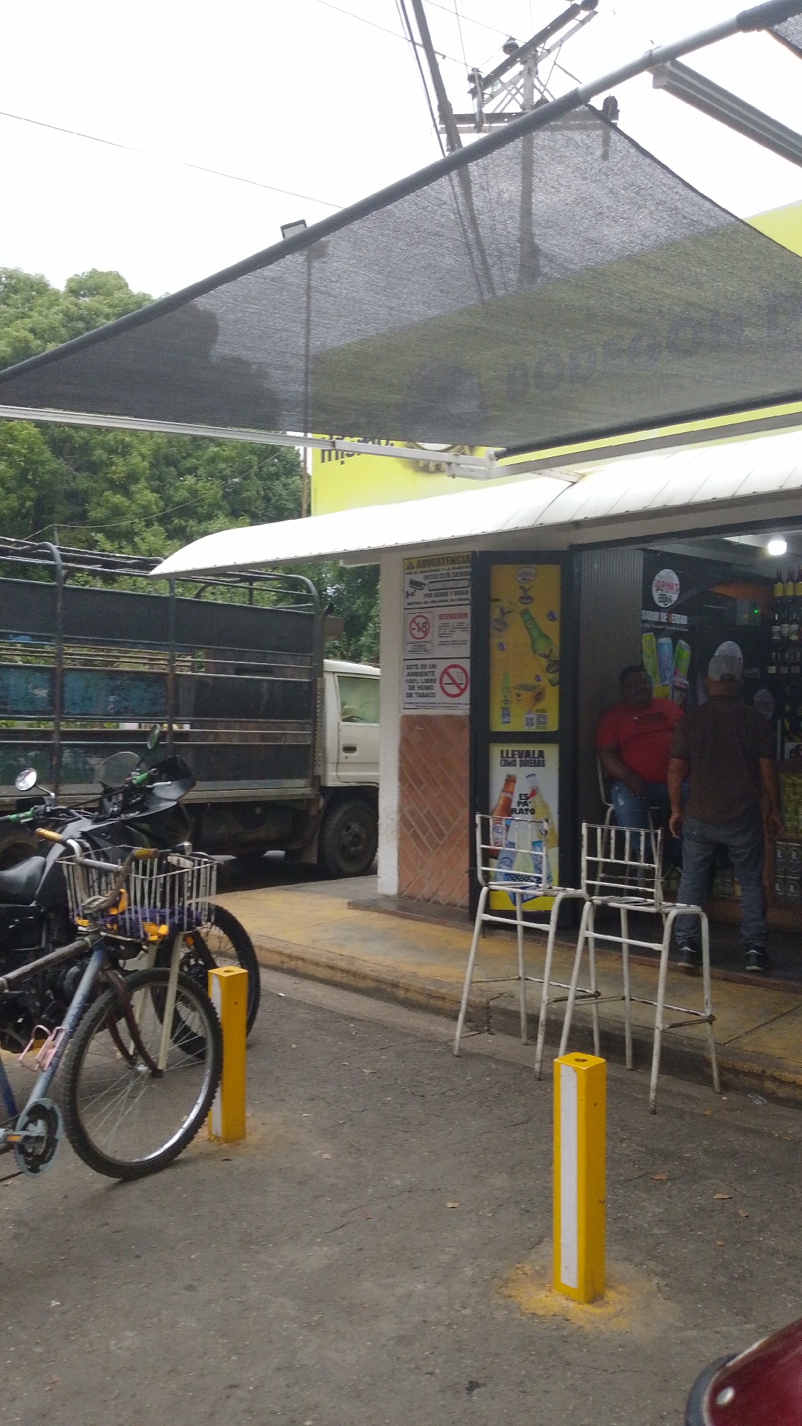 The image depicts a street scene in Tinaquillo, Venezuela. A white delivery truck with a metal cage back is parked on the street next to a storefront. A motorcycle and bicycle are parked in front of the truck. A gray shade structure extends from the storefront. The building has a white awning and advertisements for beverages. Two men are visible inside the storefront. One man, wearing a red shirt, is seated. The second man wears a brown shirt and jeans and appears to be purchasing something. Two white metal chairs sit outside the storefront. Two yellow and white striped bollards stand near the curb. The street is paved and cracked. Trees are visible in the background. The sky is overcast. A sign warns, "ESTE ES UN AMBIENTE 100% LIBRE DE HUMO DE TABACO".