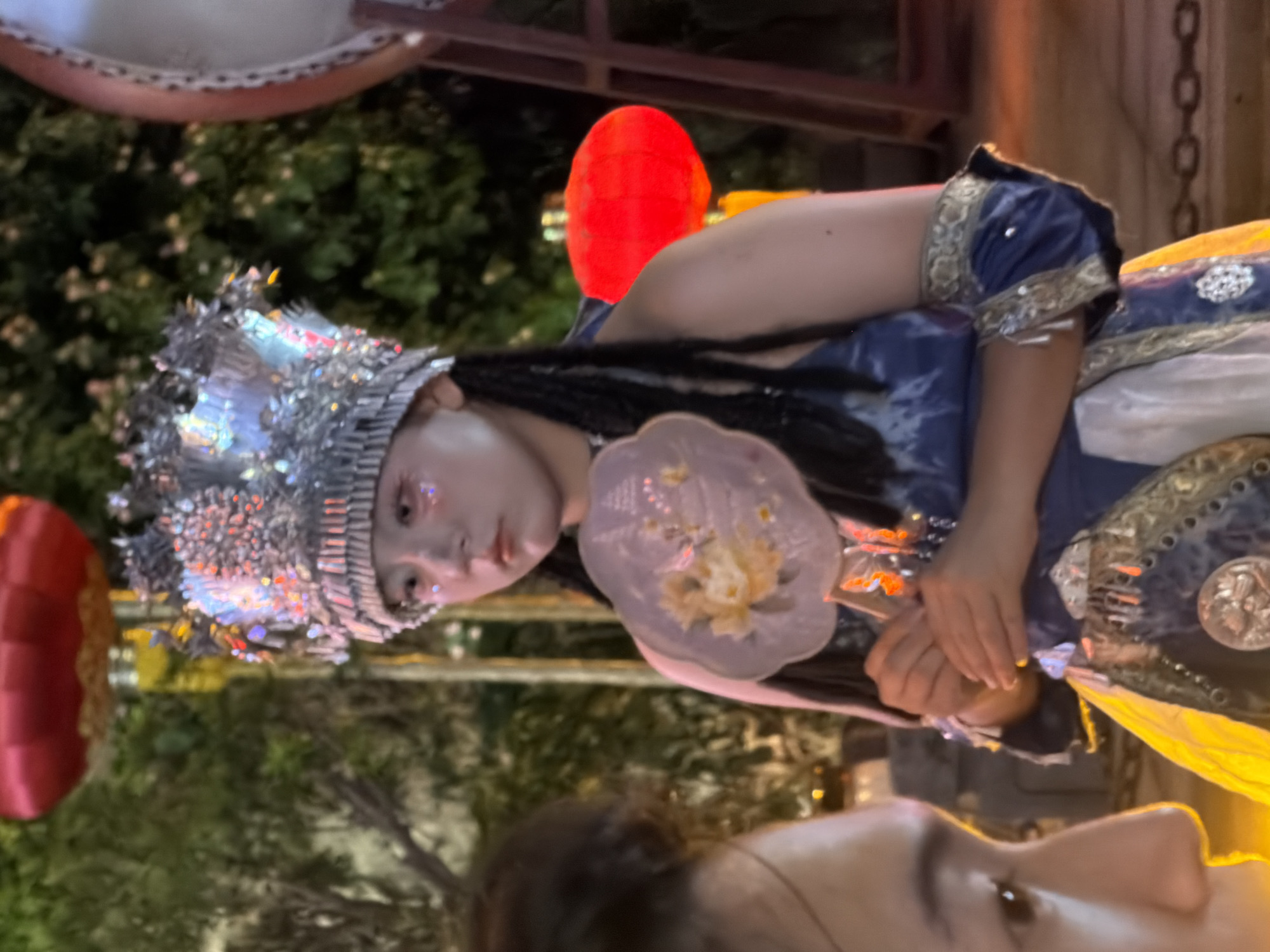 A young woman, dressed in a detailed blue and gold costume, stands in front of a crowd, holding a fan and wearing an ornate, illuminated headdress. Her face is decorated with glitter. The background suggests an outdoor nighttime event in a city, with trees, red lanterns, and a drum visible. The presence of a large drum and lanterns indicates a cultural festival or performance, possibly in Yuzhong District, China, known for its vibrant street life and cultural celebrations. The woman's attire and makeup suggest she is part of a performance or parade. The lighting is dim, illuminated by the lanterns and the woman's headdress. Another person's face is partially visible in the foreground, looking towards the woman.