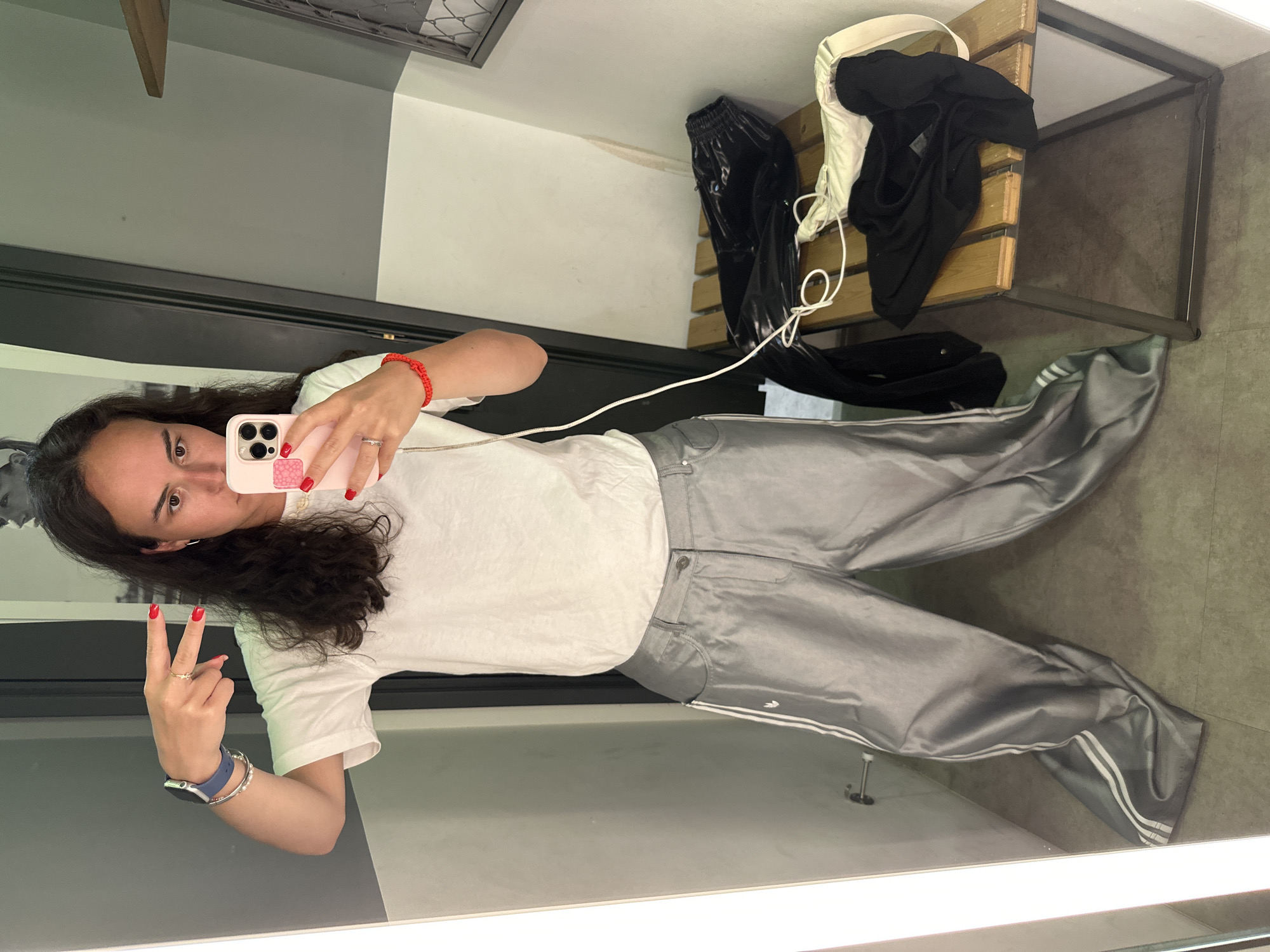 A young woman is taking a mirror selfie in a dressing room. She is wearing a white t-shirt and gray Adidas track pants. She has dark hair and is holding a pink phone. Her fingernails are painted red. She is making a peace sign with her left hand. The dressing room has a bench with some clothing on it. The location appears to be in Thailand due to the style of the room and clothing seen in the background.