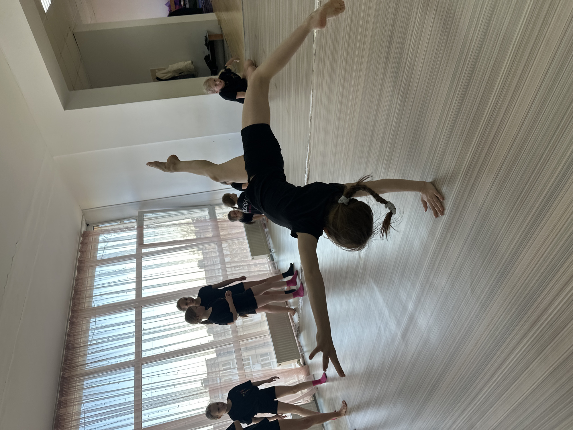 The image captures a group of young girls participating in what appears to be a dance or gymnastic class in a room featuring a large mirror. The foreground showcases a girl in a black t-shirt and black shorts, performing a handstand with her legs split. Her hair is tied back in a ponytail. Behind her, a line of girls dressed in similar black attire are visible, some wearing pink socks.
The room is well-lit, with large windows covered by light-colored curtains filling the left side of the frame. The floor has a wooden-like striped pattern. A mirror is placed on the back wall, reflecting the scene and the participants. The image's setting is inside a dance studio or gym, potentially located in Votkinsk, Russia.