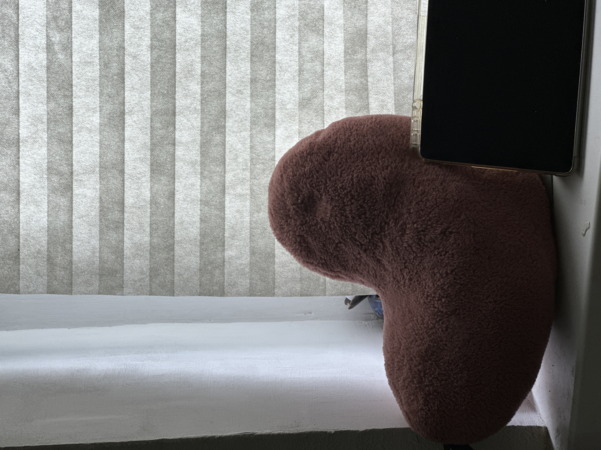 A fuzzy, heart-shaped pillow in a muted pink color rests on a windowsill. To the right of the pillow, a dark-screened tablet is propped up. Behind the pillow and tablet, a window is covered by horizontally pleated blinds. To the left of the pillow, a white sheer curtain hangs. The scene appears to be indoors.