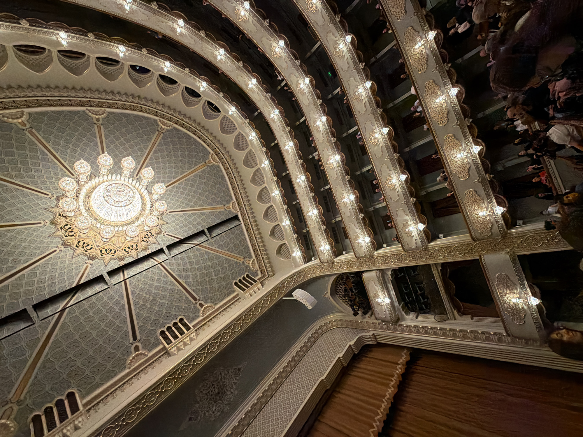 A low-angle shot captures the ornate interior of the Tbilisi Opera and Ballet State Theatre in Tbilisi, Georgia. The focal point is a large, decorative chandelier suspended from the ceiling. The ceiling is decorated with intricate patterns. Tiered balconies, complete with audience members, flank the sides of the interior space. The theater features gold-accented embellishments and architectural details. The stage is partially visible.
