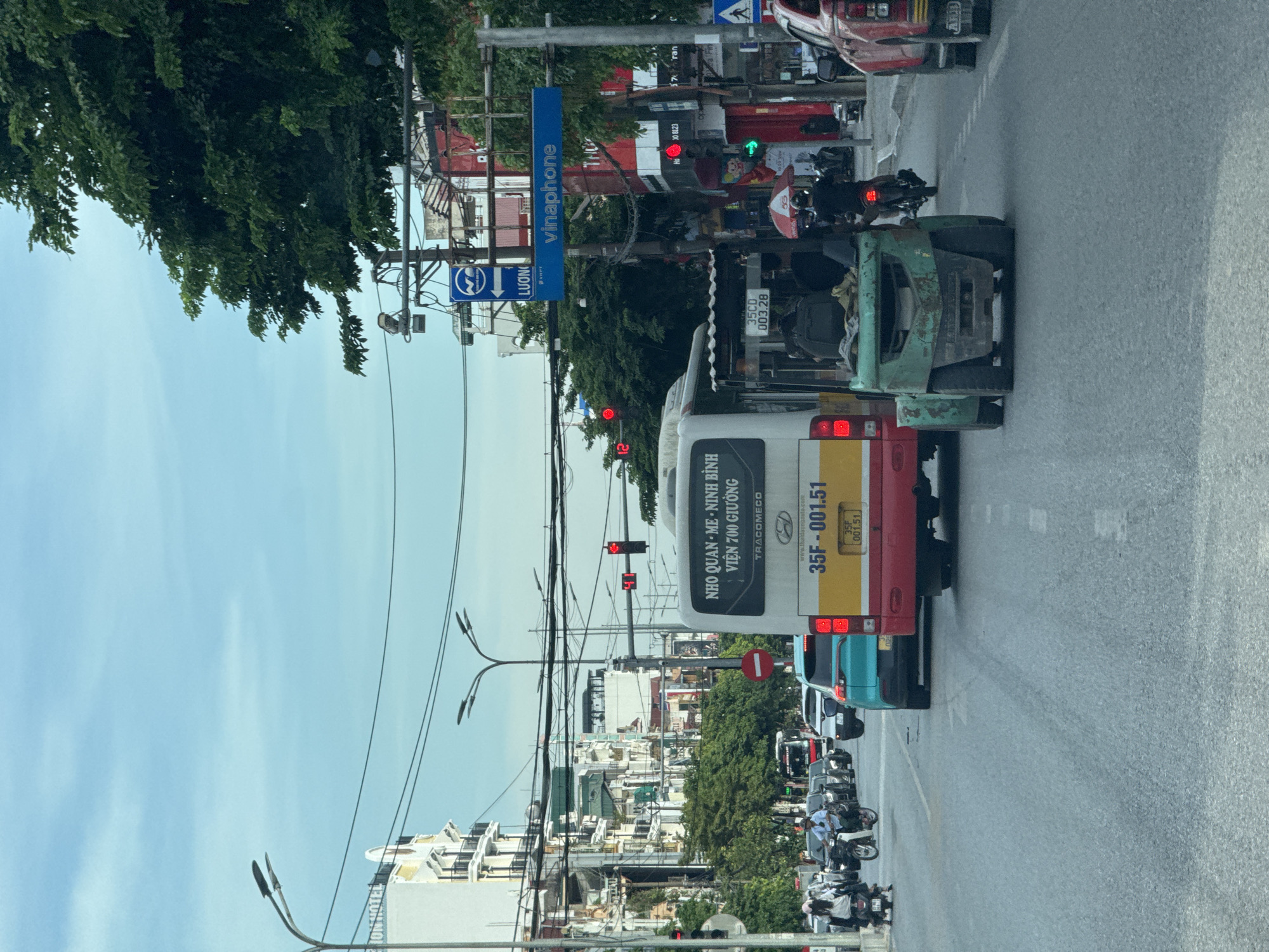 The image captures a street scene in Hoa Lư Ward, Vietnam, under an overcast sky. A red and yellow bus, labeled "NHO QUAN - ME - NINH BINH, VIEN 700 GIUONG" and with license plate "35F - 001.51," is prominent in the foreground, stopped at what appears to be a traffic signal. To its right, a green forklift is also stopped, with two individuals visible inside.
A turquoise car is positioned behind the bus, and numerous other vehicles, including cars and motorcycles, are present on the multi-lane road. Several traffic lights are visible, with at least one displaying red. Overhead, power lines crisscross the sky, and streetlights are mounted on poles. Buildings line the street, some appearing to be hotels or commercial establishments, with visible signage including "VINAPHONE." Trees and foliage are present, contributing to the urban landscape. People are seen on motorcycles and walking, suggesting normal daily activity. The road surface is asphalt.