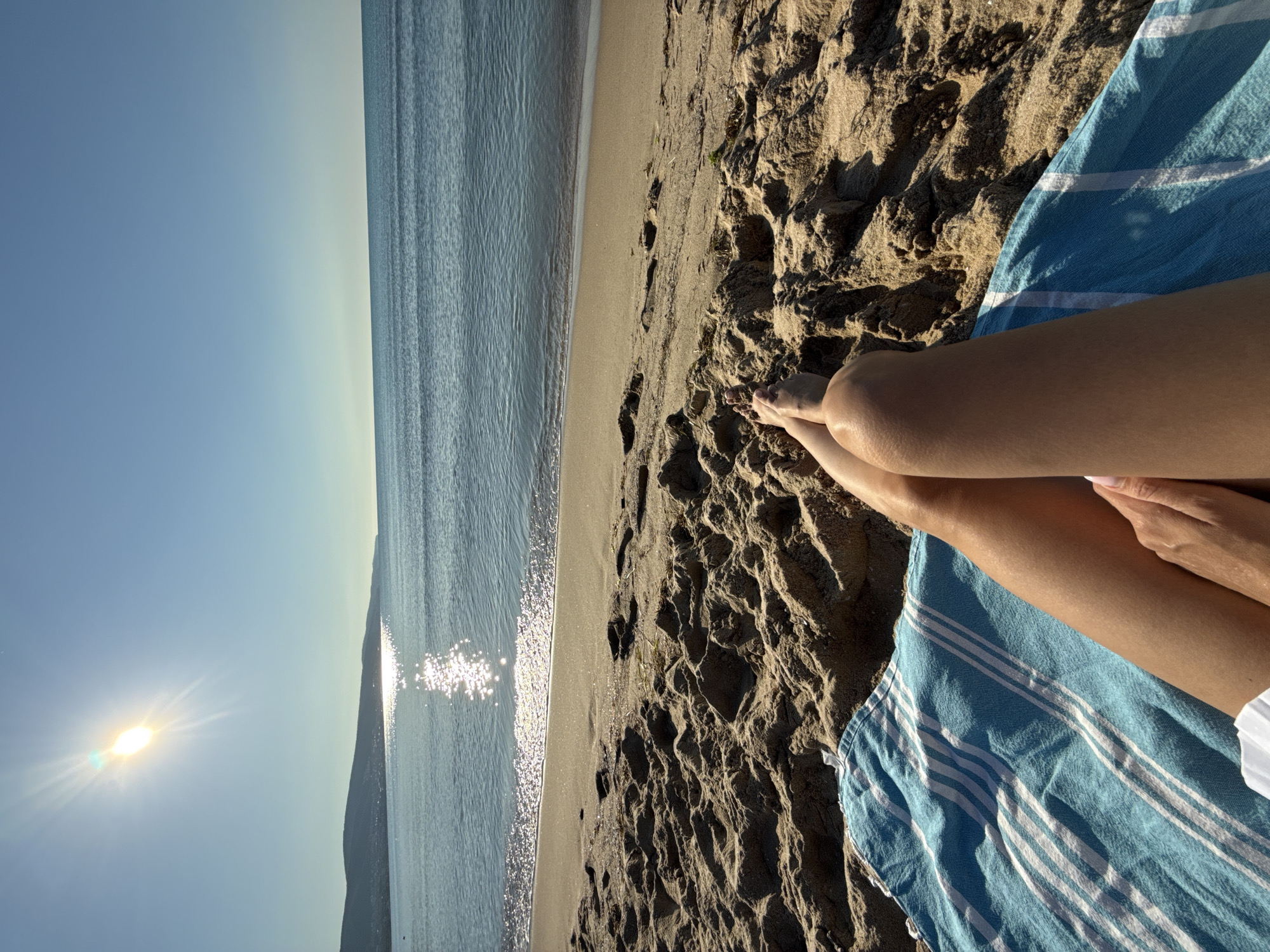 A person's legs and feet are visible, resting on a blue and white striped towel on a sandy beach. The sun shines brightly in the clear blue sky, casting a reflection on the calm sea. In the distance, a coastline with hills and buildings can be seen. The image captures a peaceful moment at the beach in Nesebar, Bulgaria.