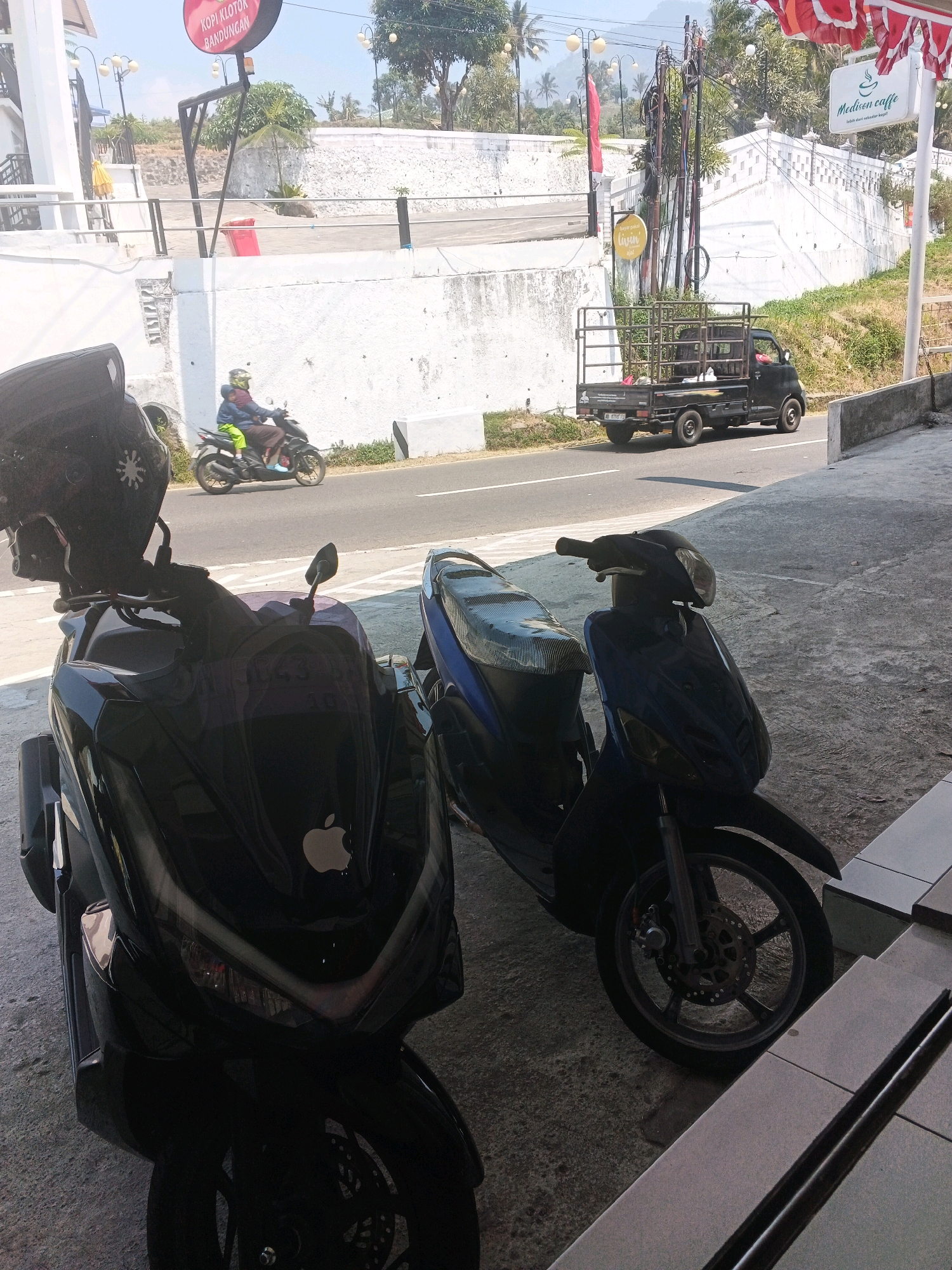 The image shows a street scene in Pakopen, Indonesia, with a focus on two motorcycles parked on a paved area. In the foreground, a black scooter is prominently displayed, featuring a stylized Apple logo on its front. To its right, a blue and black scooter is also parked. In the background, a person wearing a helmet rides a motorcycle with a passenger. A black pickup truck is visible driving on the road, with a white building and lush greenery in the distance. Signs for "Kopi Klotok Bandungan" and "Medicon Caffe" are also visible, indicating the presence of local businesses. The overall environment appears to be a sunny day with clear skies.