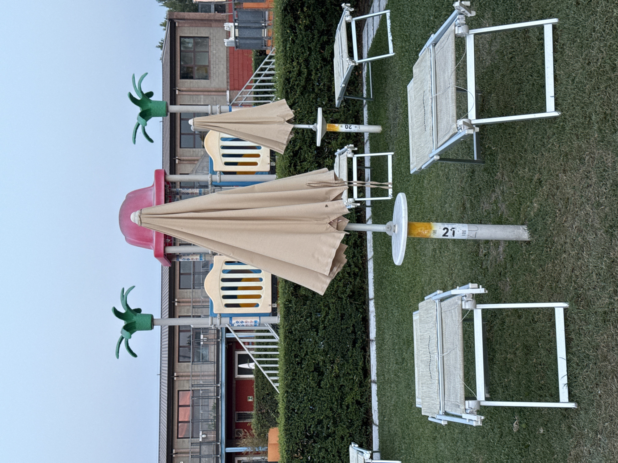 The image depicts a playground area, likely at a resort or public facility, with a clear focus on the foreground and middle ground. In the foreground, there are several folded-up lounge chairs and a few partially open lounge chairs on a grassy lawn. Two large, tan umbrellas are prominently displayed, one in the center and another to the right, both in a partially closed or collapsed state. A pole with a yellow band and the number "21" is visible between the lounge chairs.
The background reveals a playground structure with slides and climbing elements, painted in bright colors like red, yellow, and blue. Two stylized green palm tree-shaped decorations are positioned on top of poles, framing the playground. Behind the playground, a multi-story building with a grey roof and a façade featuring windows and balconies is visible. The overall scene suggests an outdoor recreational space, possibly a beach resort or a park, set against a muted, overcast sky. There is no indication of the city of Parma, Italy, in the image itself, nor are there any people present.
